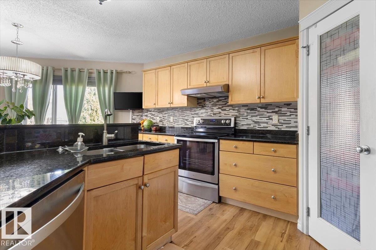 Kitchen featuring wood cabinetry, dark countertops, a mosaic tile backsplash, stainless steel appliances, and wood-finish flooring - 3356 25 Ave, Edmonton, AB - Indoor Photo Showing Kitchen With Double Sink