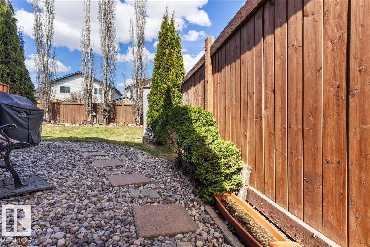 Landscaped backyard featuring a wood fence, gravel patio with stepping stones, manicured lawn, evergreen shrubs, and tall deciduous trees - 3356 25 Ave, Edmonton, AB - Outdoor