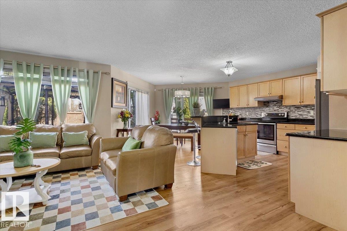 Open-concept living area featuring wood-finish flooring, a kitchen with light wood cabinetry and dark countertops, stainless steel appliances, and a tiled backsplash - 3356 25 Ave, Edmonton, AB - Indoor