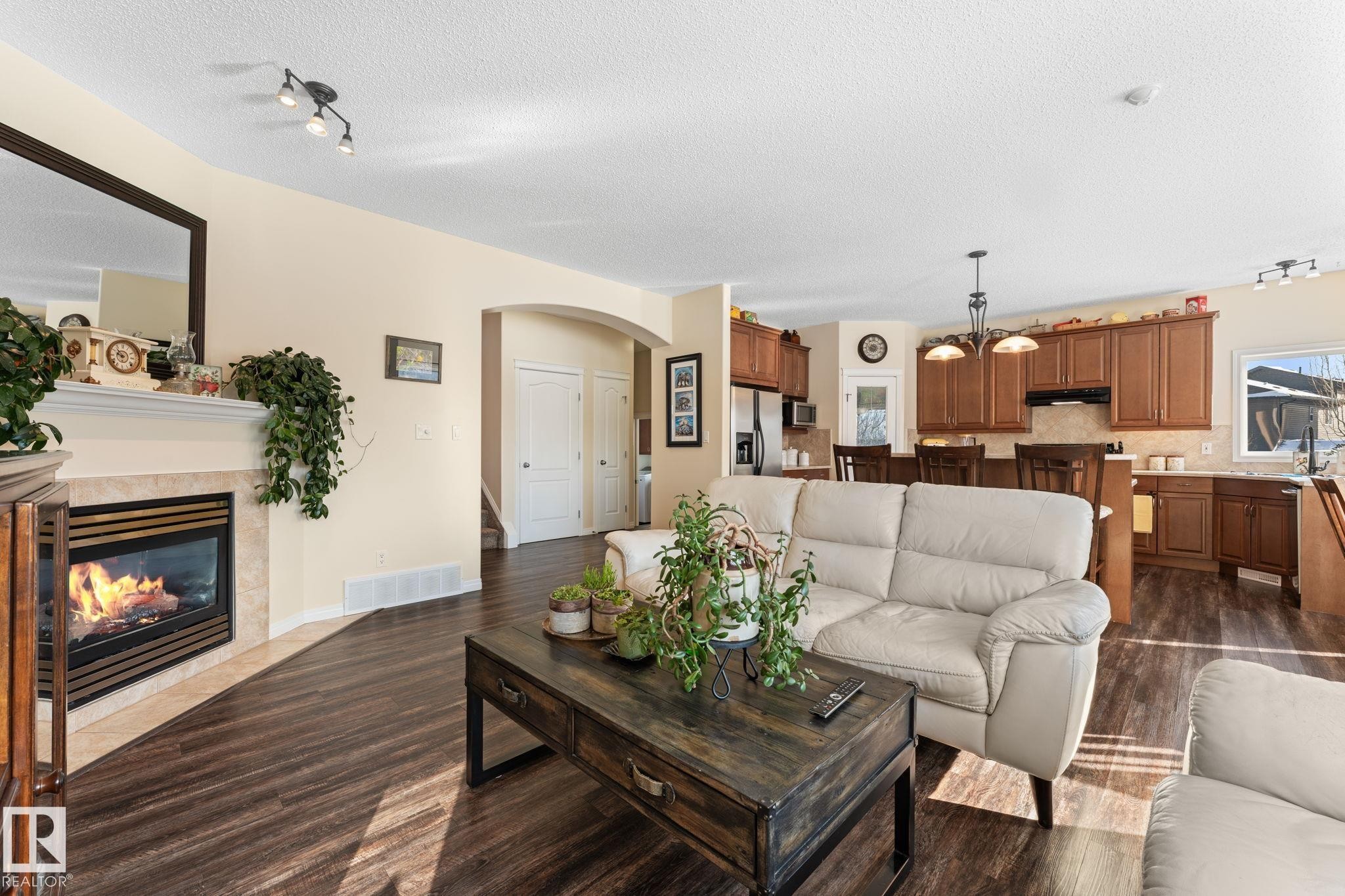 Open-concept living space with dark wood-finish flooring, a built-in fireplace featuring a tile surround and mantel, and an arched entry - 112 Lakeland Point(E), Beaumont, AB - Indoor Photo Showing Living Room With Fireplace