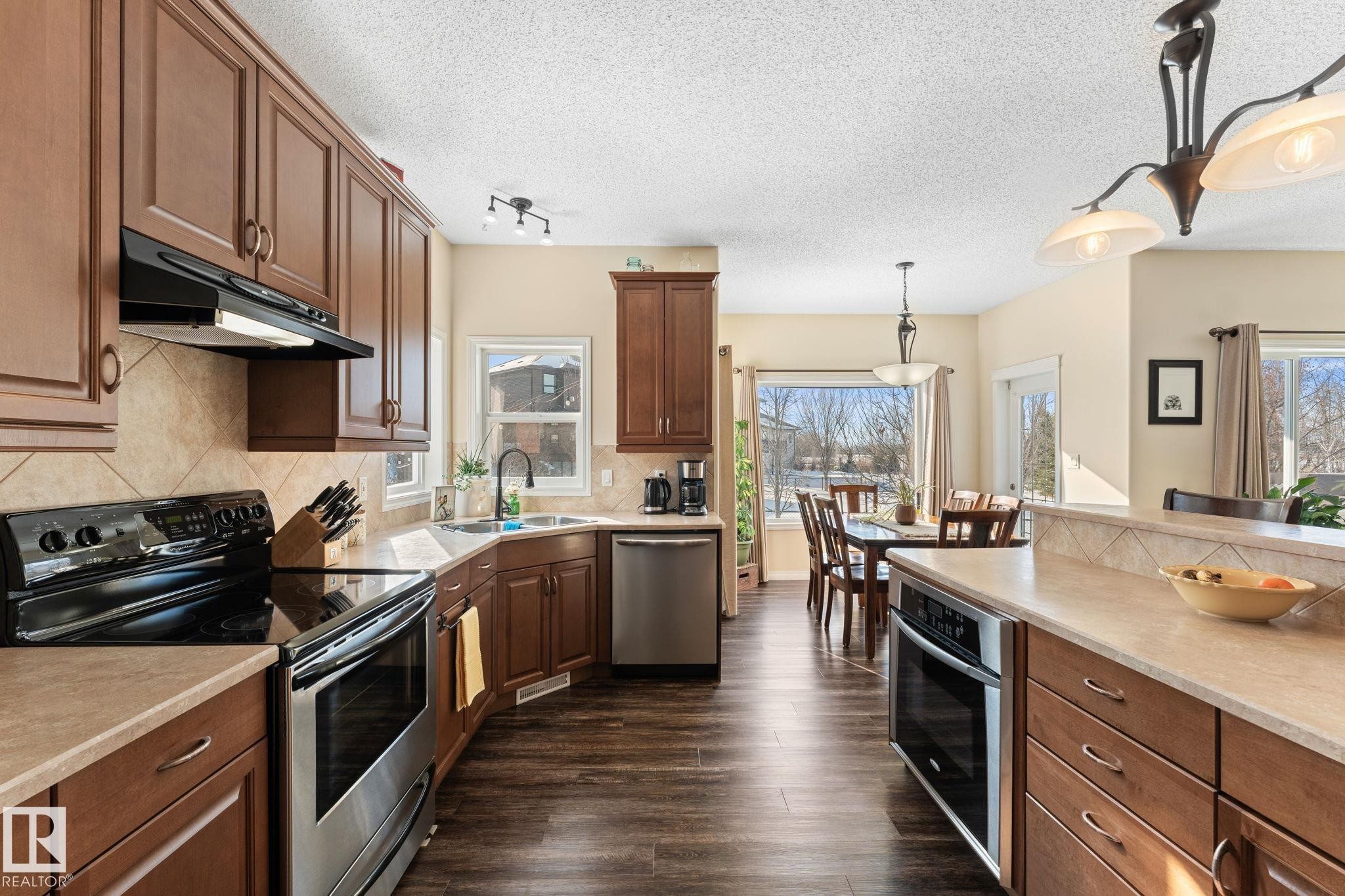 Well-appointed kitchen featuring wood-finish flooring, warm-toned cabinetry, light countertops, stainless steel appliances, and a tiled backsplash - 112 Lakeland Point(E), Beaumont, AB - Indoor Photo Showing Kitchen With Double Sink With Upgraded Kitchen
