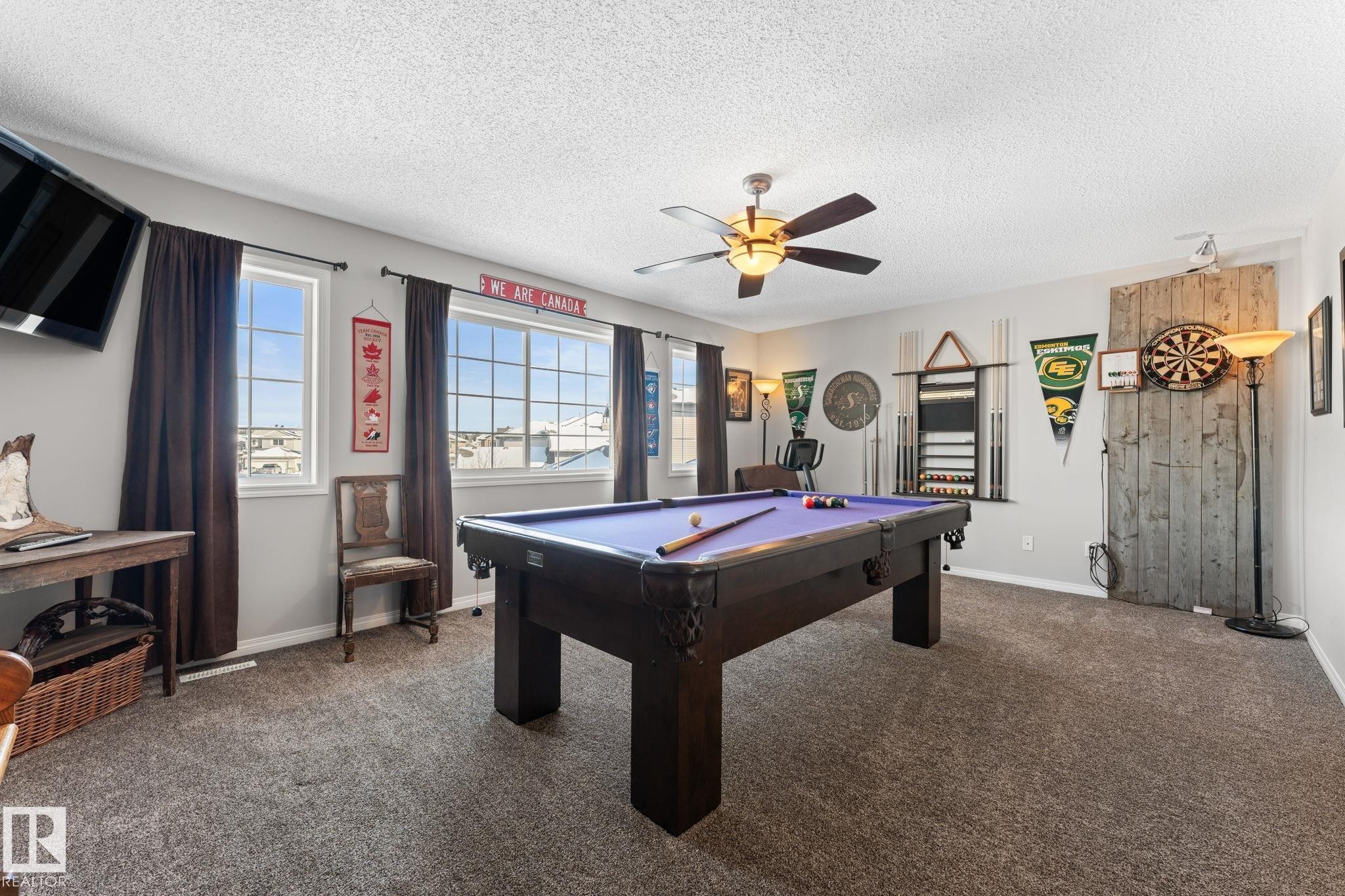 Spacious recreation room featuring a built-in pool table, broad windows, textured carpeting, a ceiling fan with integrated lighting, and a prominent wood-panel accent wall - 112 Lakeland Point(E), Beaumont, AB - Indoor Photo Showing Other Room