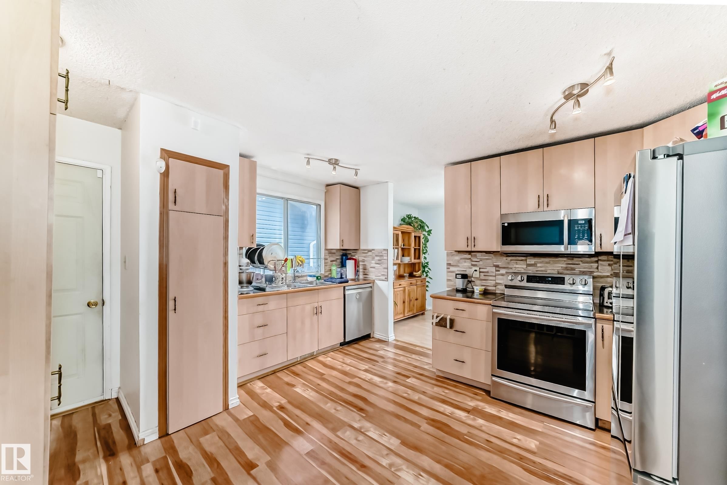 Kitchen featuring wood-finish flooring, light wood cabinetry, stone-look backsplash, and stainless steel appliances - 1804 51 Street Nw, Edmonton, AB - Indoor Photo Showing Kitchen With Double Sink