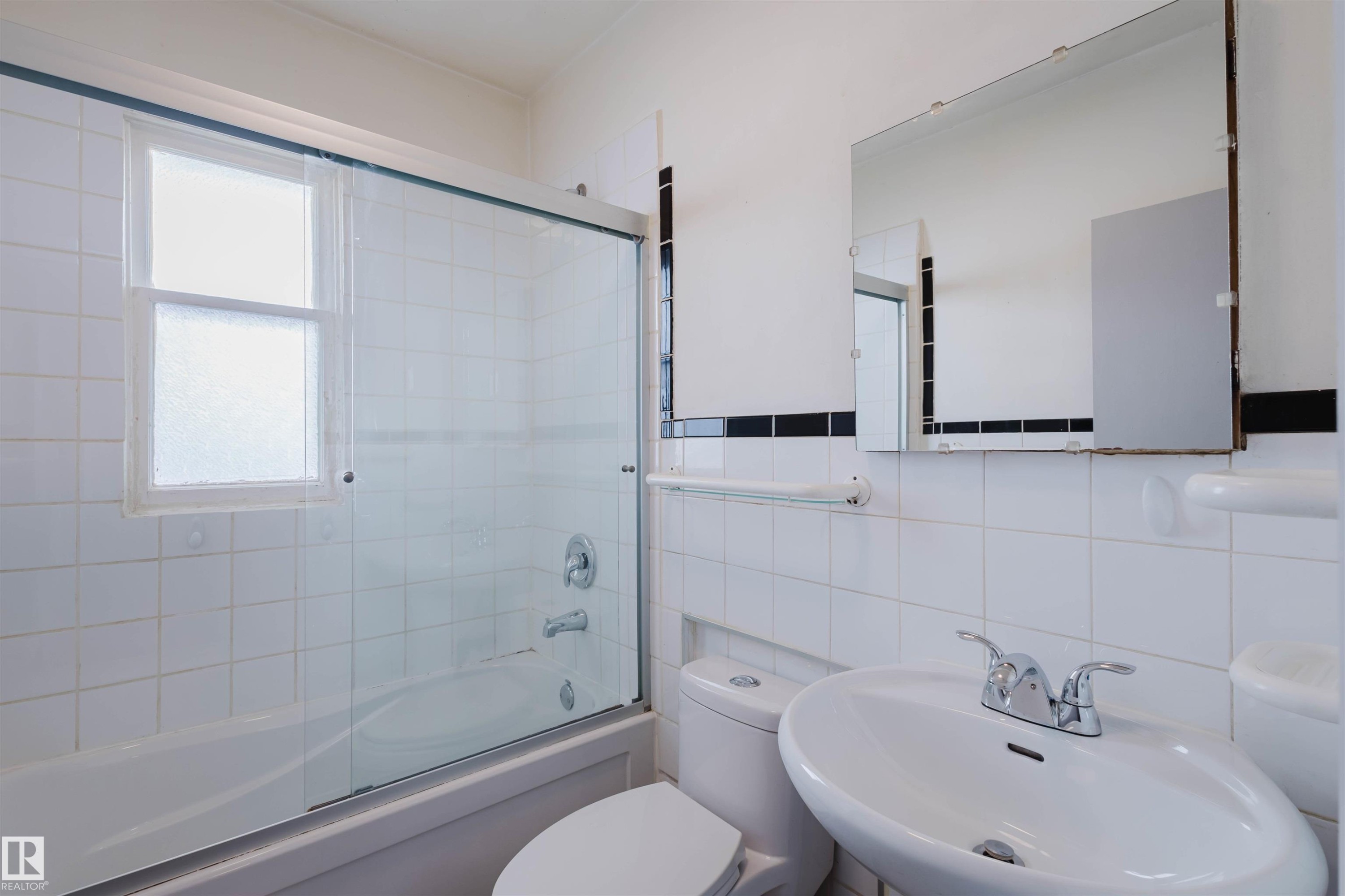 Bathroom featuring a white ceramic tile surround, a single-pane window with frosted glass, a chrome faucet and showerhead, a sliding glass shower door, and a wall-mounted rectangular mirror - 10511 80 Street, Edmonton, AB - Indoor Photo Showing Bathroom