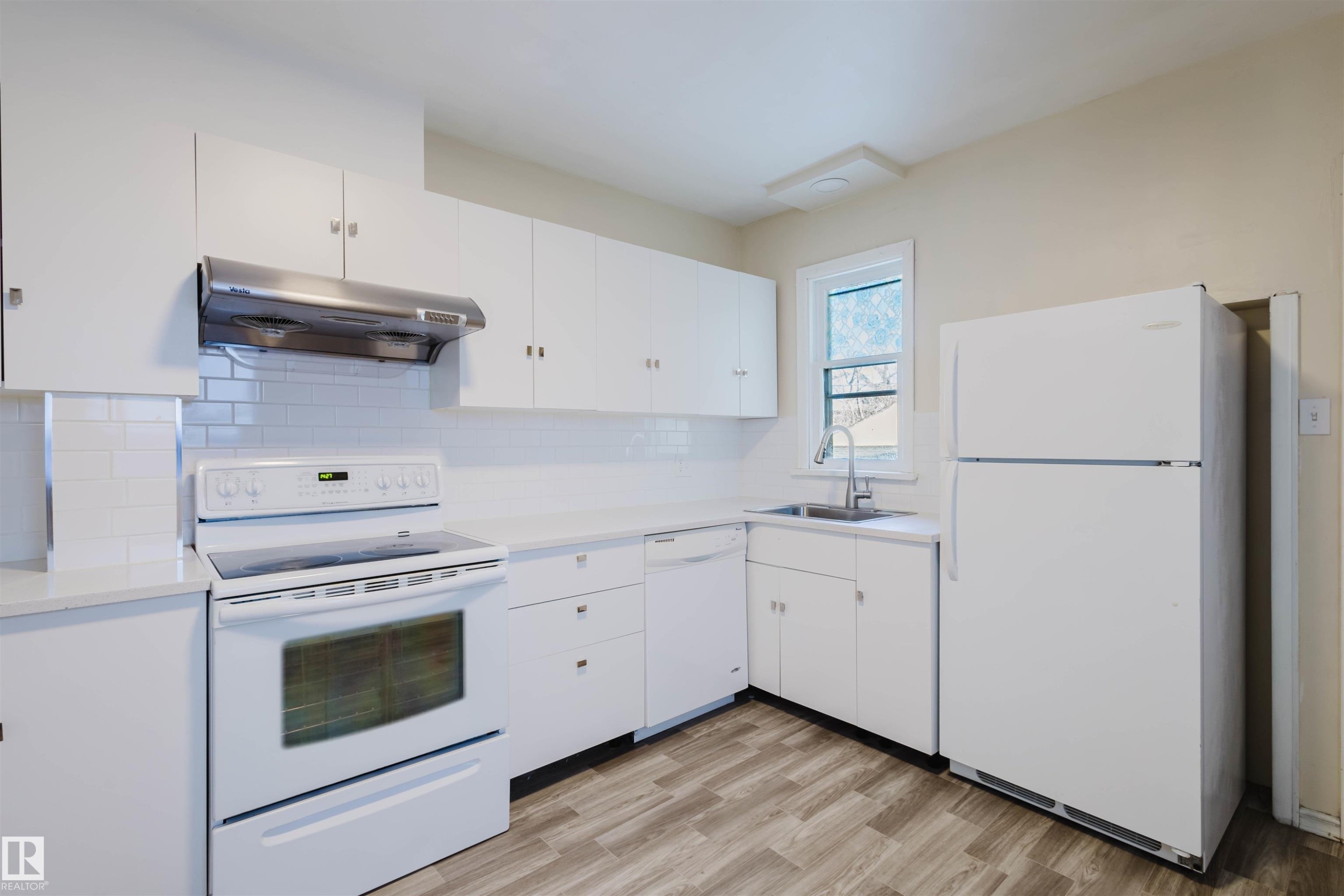 Kitchen featuring white cabinetry, white subway tile backsplash, light-toned countertops, wood-finish flooring, and a stainless steel range hood - 10511 80 Street, Edmonton, AB - Indoor Photo Showing Kitchen