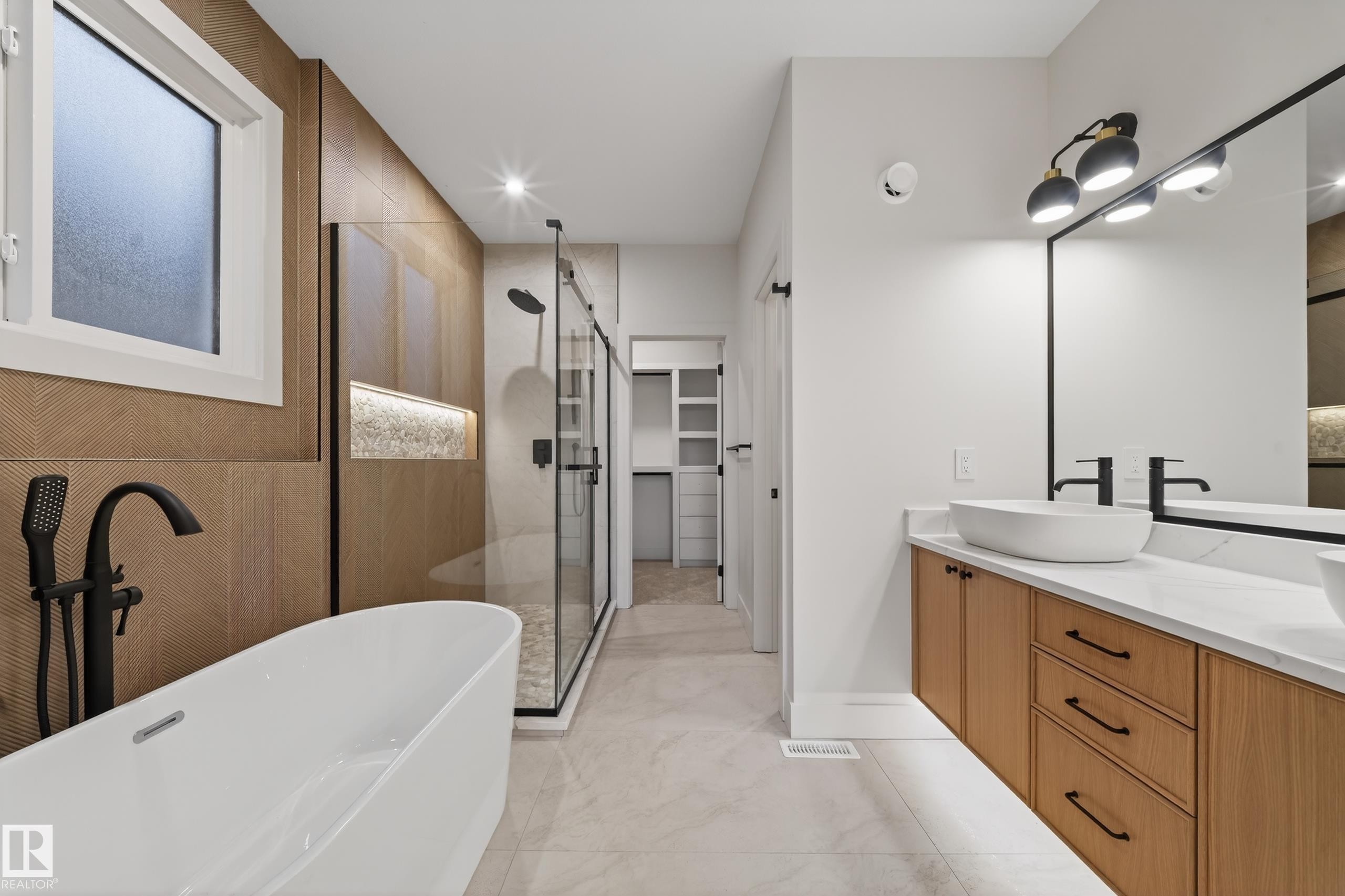 Spacious bathroom featuring a freestanding soaking tub with matte black fixtures, a walk-in shower with a glass enclosure, a dual vanity with vessel sinks and quartz countertops, wood-finish cabinetry, and textured tile accents - 4276 Charles Close, Edmonton, AB - Indoor Photo Showing Bathroom