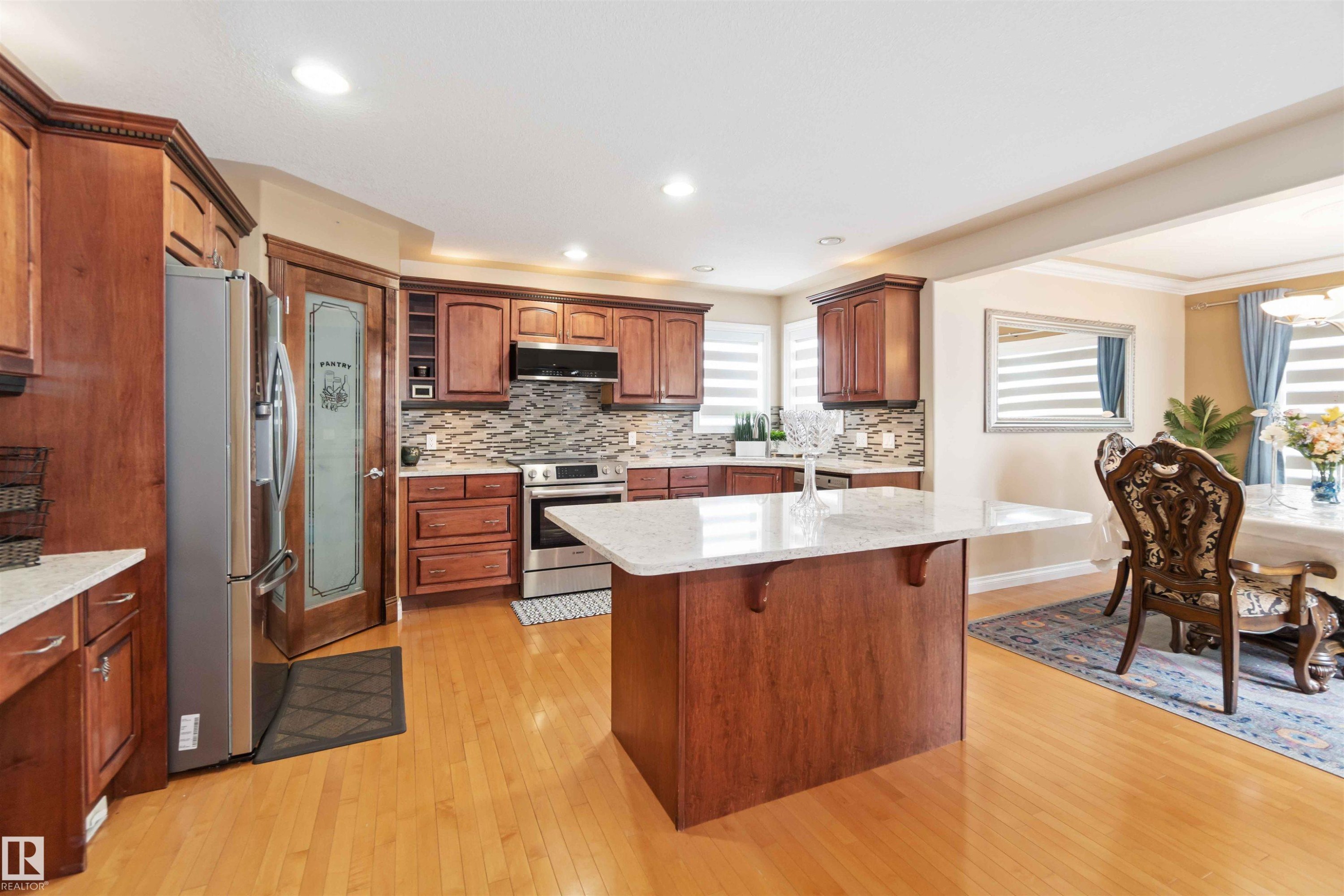 Kitchen featuring wood-finish cabinetry, stainless steel appliances, a center island with a light-toned countertop, a tile backsplash, and light wood-finish flooring - 6116 53 Avenue, Beaumont, AB - Indoor Photo Showing Kitchen With Stainless Steel Kitchen With Upgraded Kitchen