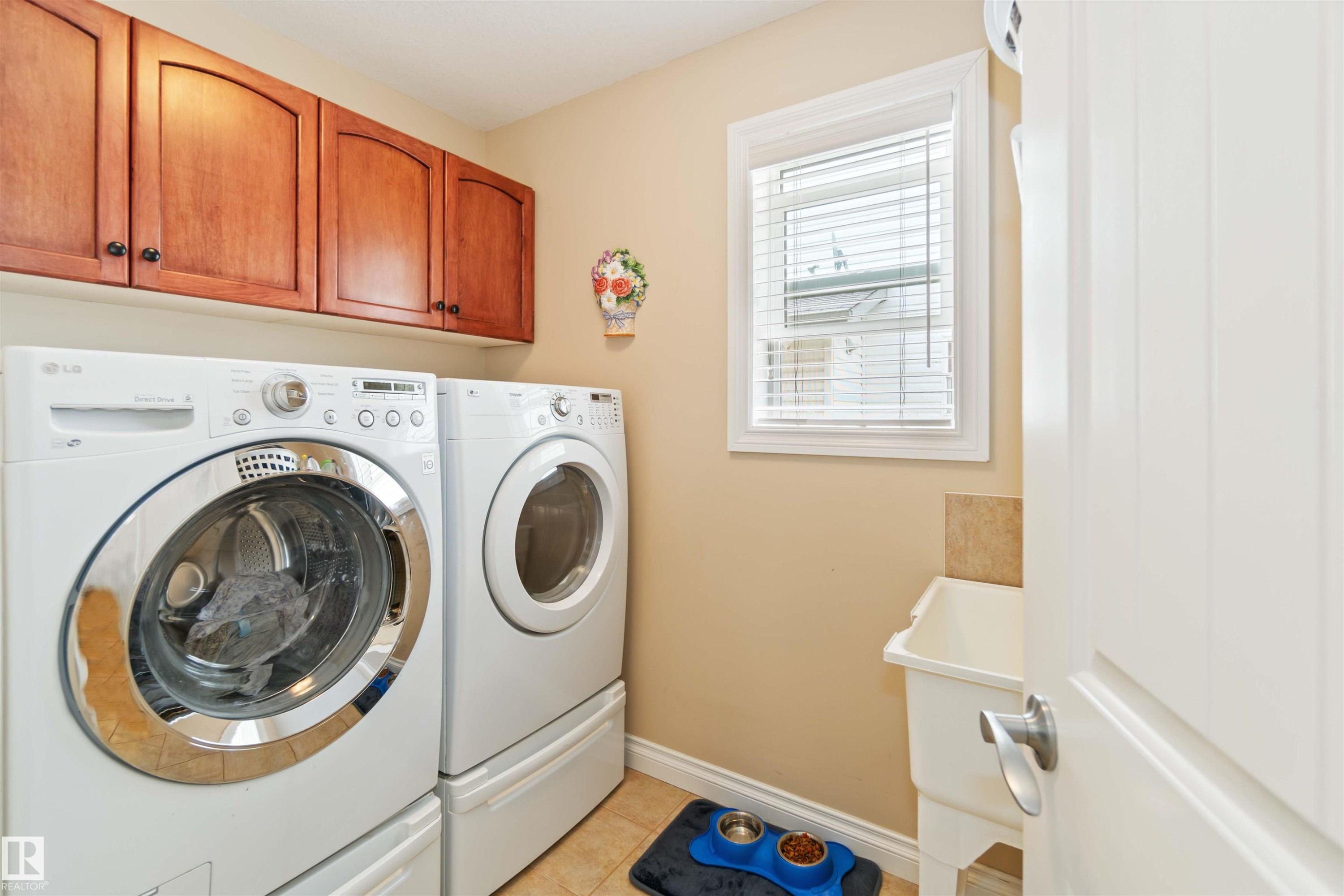 Dedicated laundry room featuring ceramic tile flooring, wood cabinetry, a utility sink with a tiled backsplash, and a window with blinds - 6116 53 Avenue, Beaumont, AB - Indoor Photo Showing Laundry Room