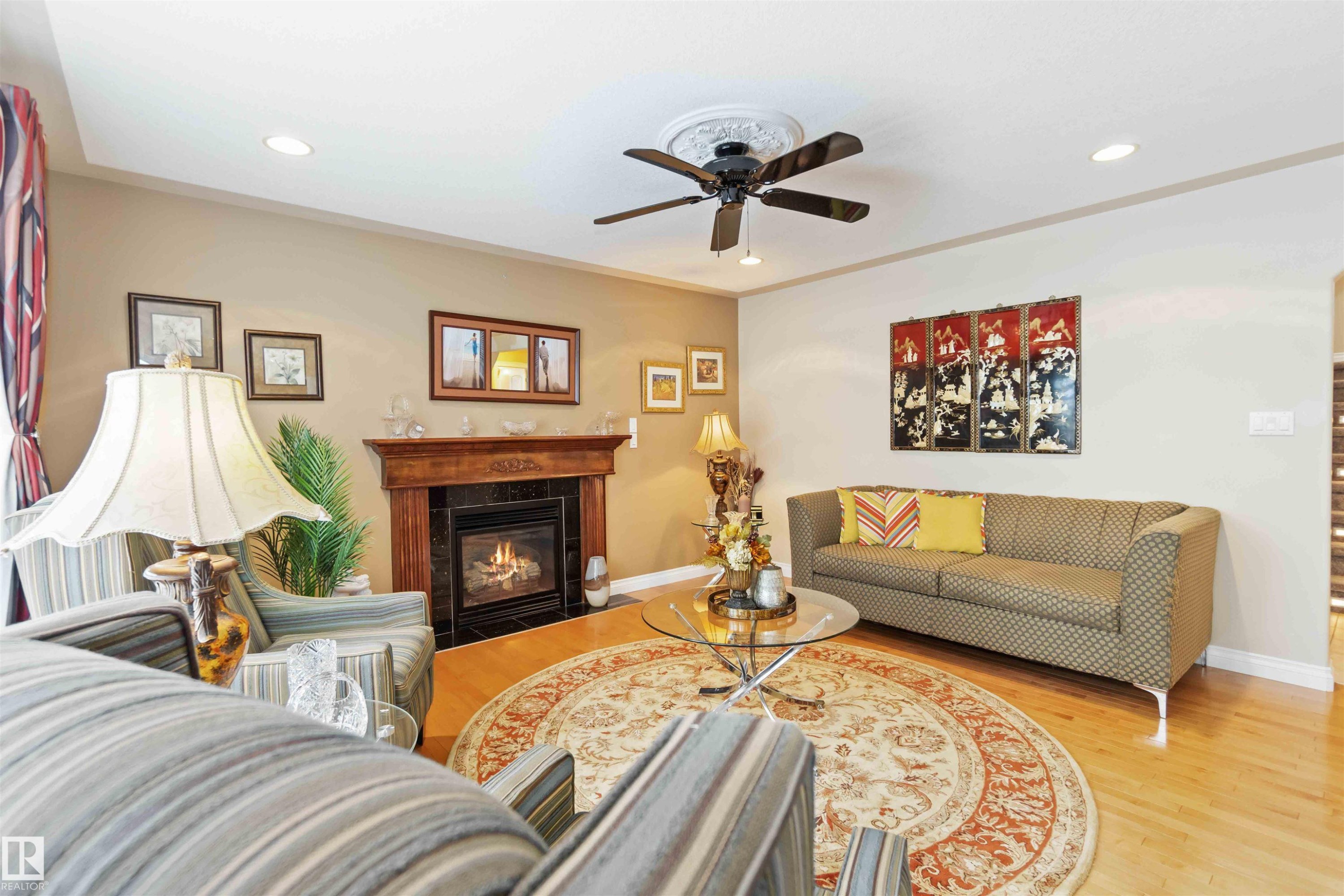 Living area featuring wood-finish flooring, a fireplace with a wood mantle and dark stone surround, and a ceiling fan with integrated lighting - 6116 53 Avenue, Beaumont, AB - Indoor Photo Showing Living Room With Fireplace