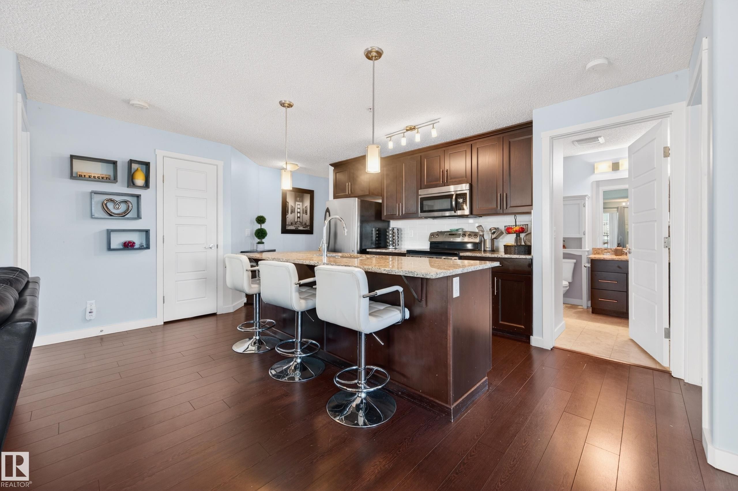 Open-concept kitchen featuring dark wood cabinetry, stainless steel appliances, and a central island with a light-toned countertop - 340 6076 Schonsee Way, Edmonton, AB - Indoor Photo Showing Kitchen With Stainless Steel Kitchen With Upgraded Kitchen