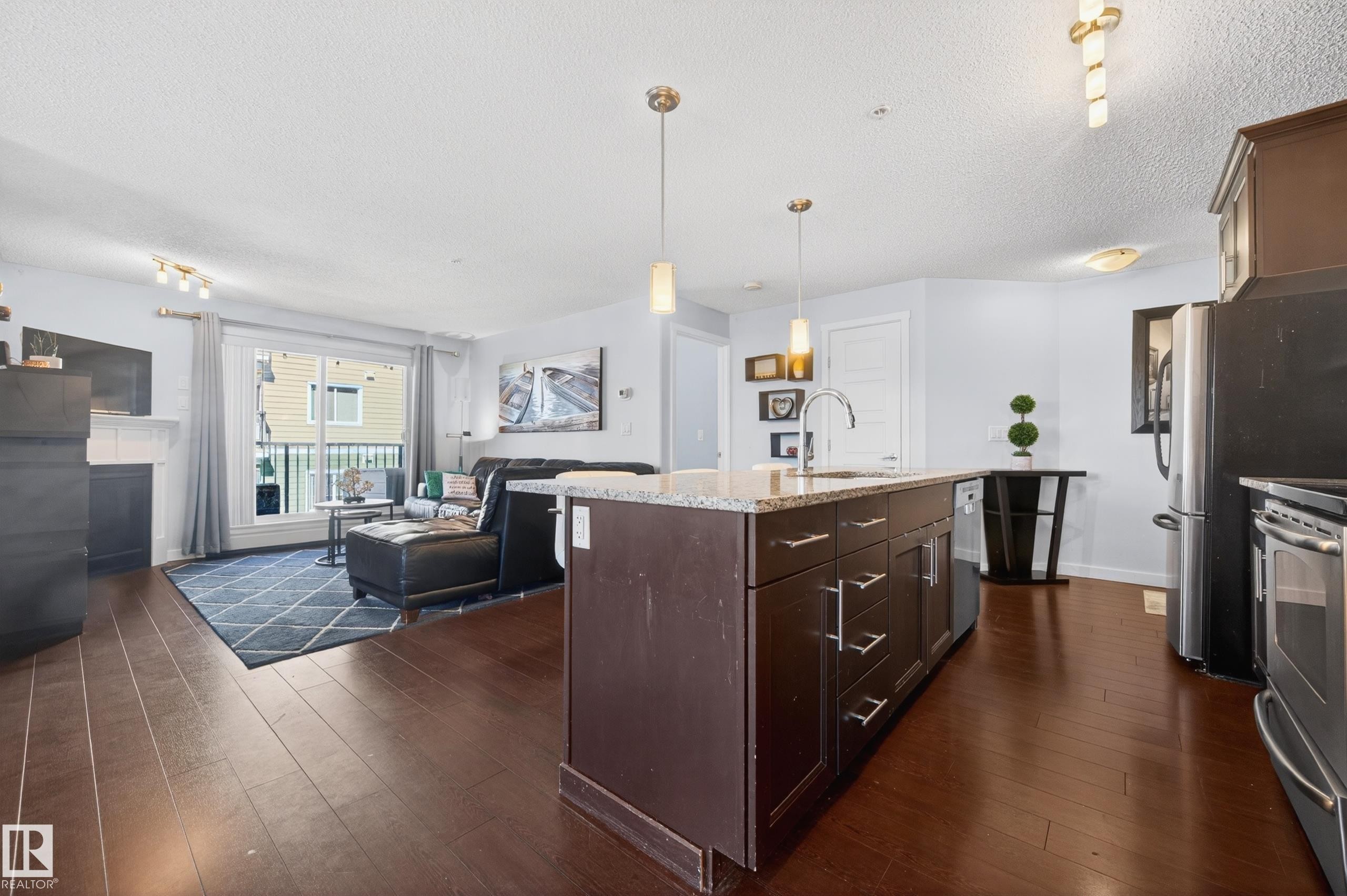 Open-concept living area featuring a kitchen island with stone-finish countertop, dark wood-finish cabinetry, stainless steel appliances, and wood-finish flooring - 340 6076 Schonsee Way, Edmonton, AB - Indoor Photo Showing Kitchen With Upgraded Kitchen