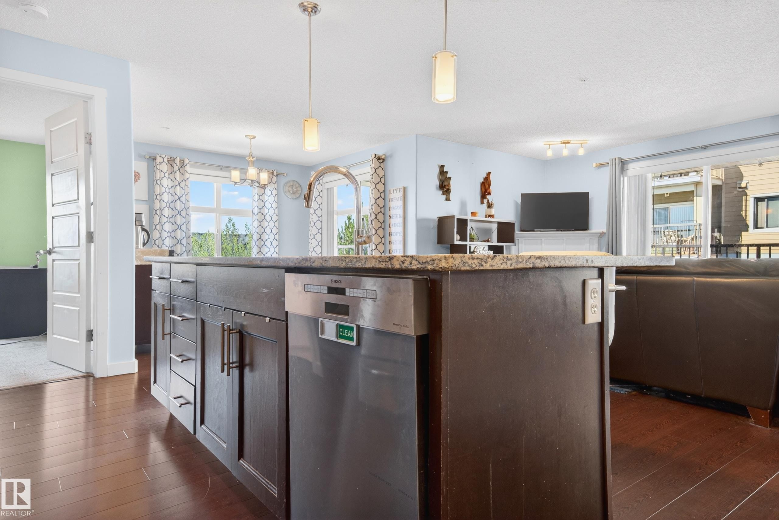 Kitchen island featuring dark wood cabinetry, stainless steel dishwasher, and a stone countertop - 340 6076 Schonsee Way, Edmonton, AB - Indoor Photo Showing Kitchen With Upgraded Kitchen