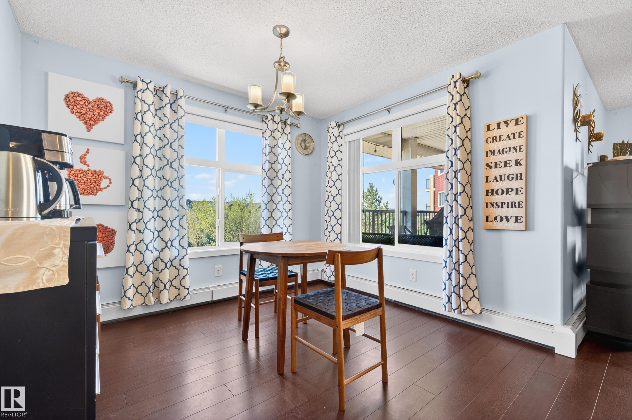 Wood-finish flooring provides a rich foundation for this bright dining area, featuring two large windows with patterned drapes - 340 6076 Schonsee Way, Edmonton, AB - Indoor Photo Showing Dining Room