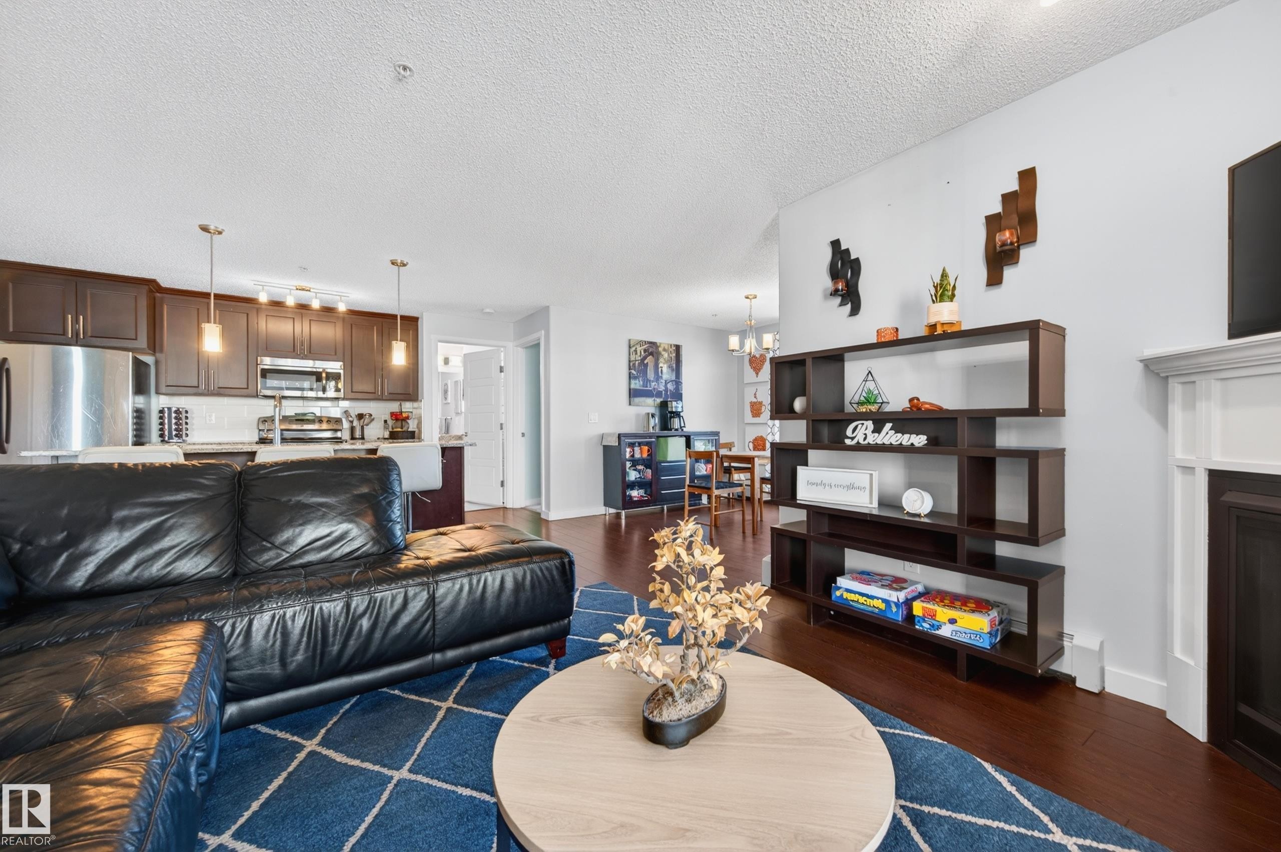 Open-concept living area featuring rich wood-finish flooring, a contemporary built-in shelving unit, and a kitchen with dark cabinetry and stainless steel appliances - 340 6076 Schonsee Way, Edmonton, AB - Indoor Photo Showing Living Room
