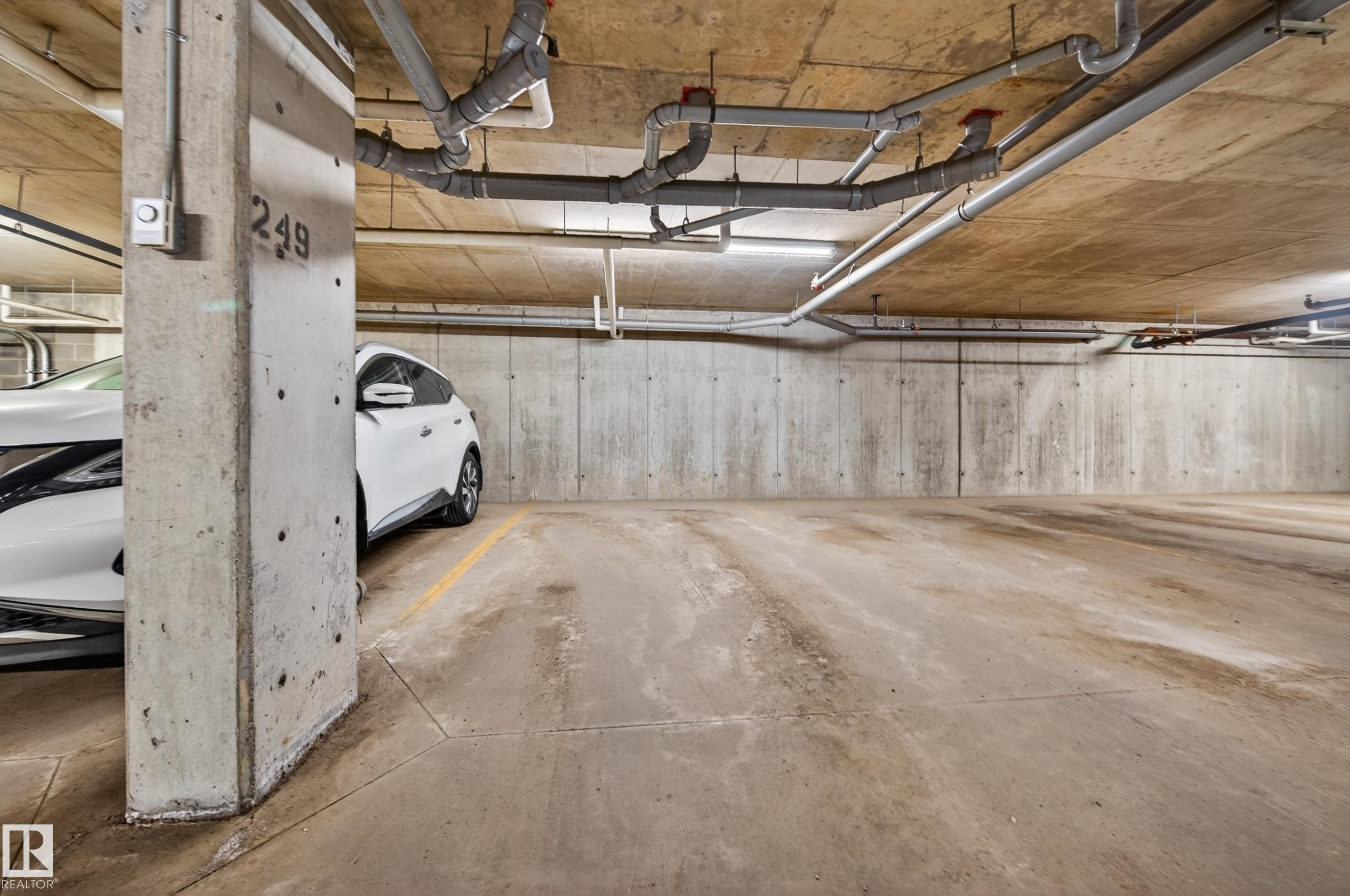 Concrete parking structure featuring exposed concrete walls and ceiling, overhead utility piping, fluorescent lighting, and a marked concrete parking space - 340 6076 Schonsee Way, Edmonton, AB - Indoor Photo Showing Garage