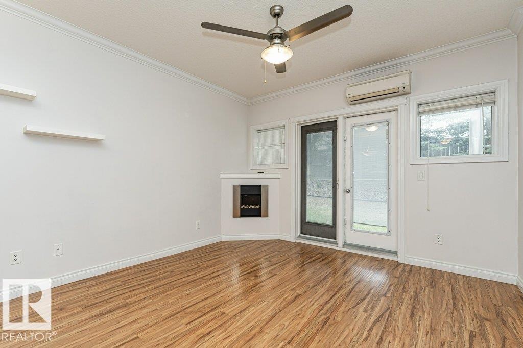 Spacious room featuring wood-finish flooring, a built-in fireplace, a ceiling fan, and multiple windows - #145 10121 80 Av Nw, Edmonton, AB - Indoor Photo Showing Other Room With Fireplace