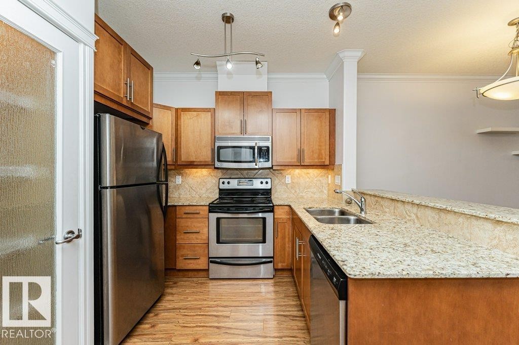 Kitchen featuring wood-finish flooring, stainless steel appliances, light wood cabinetry, and a double basin sink - #145 10121 80 Av Nw, Edmonton, AB - Indoor Photo Showing Kitchen With Stainless Steel Kitchen With Double Sink