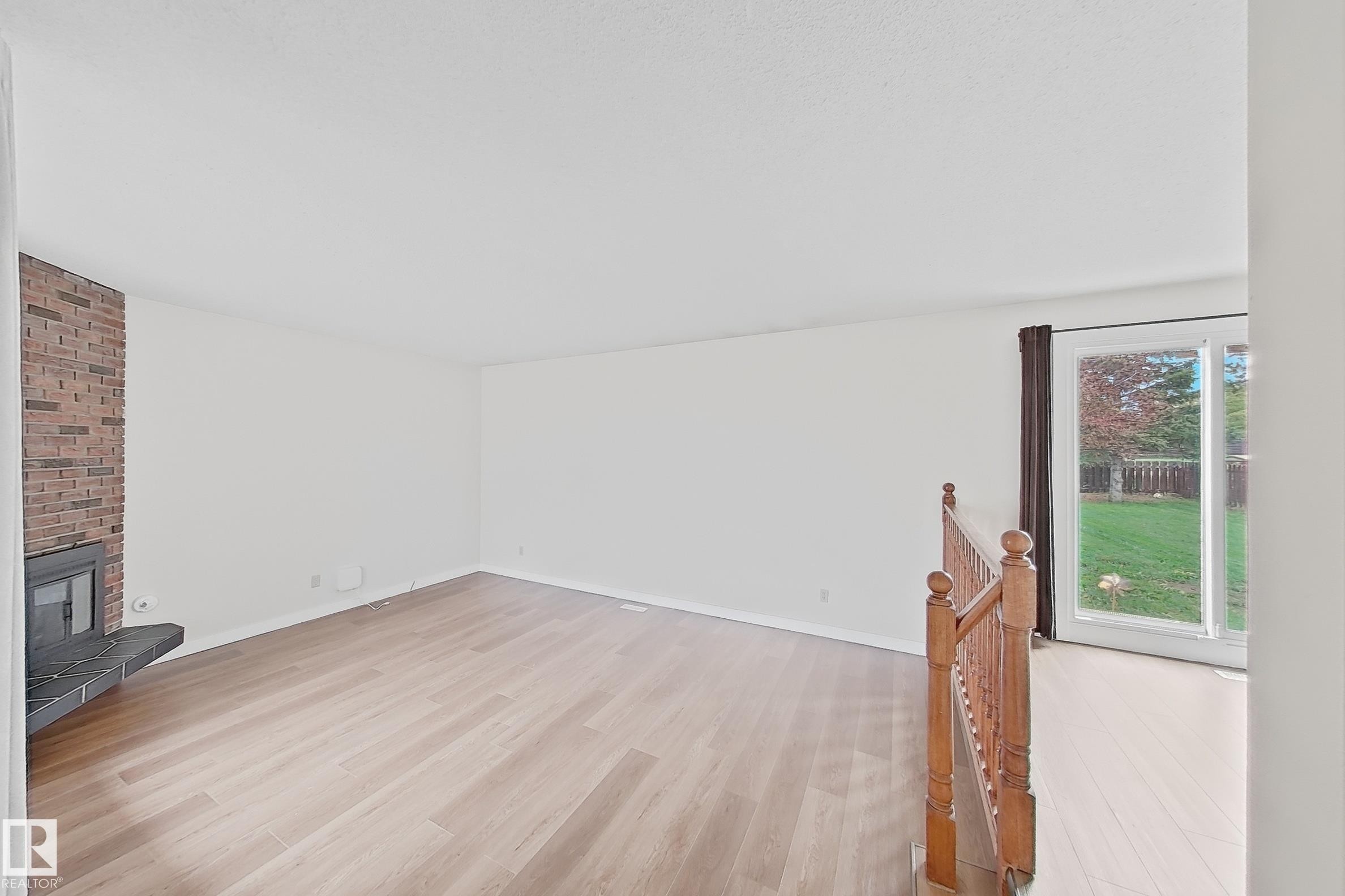 Spacious room featuring light wood-finish flooring, a brick-faced fireplace with a dark hearth, and a sliding glass door providing access to the backyard - 1731 51 Street, Edmonton, AB - Indoor Photo Showing Other Room With Fireplace