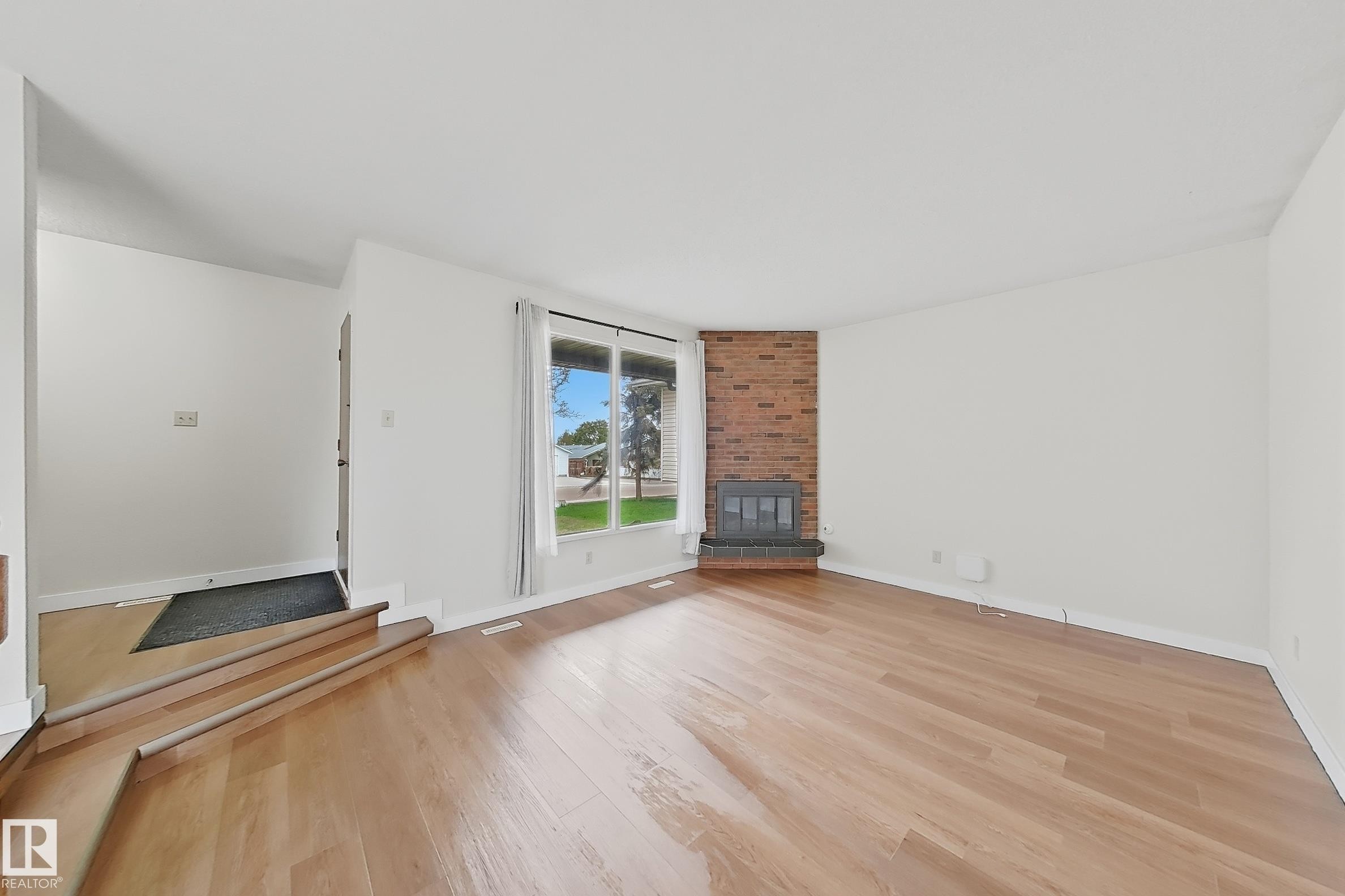 Spacious living area featuring wood-finish flooring, a prominent brick fireplace with hearth, and large windows providing ample natural light - 1731 51 Street, Edmonton, AB - Indoor