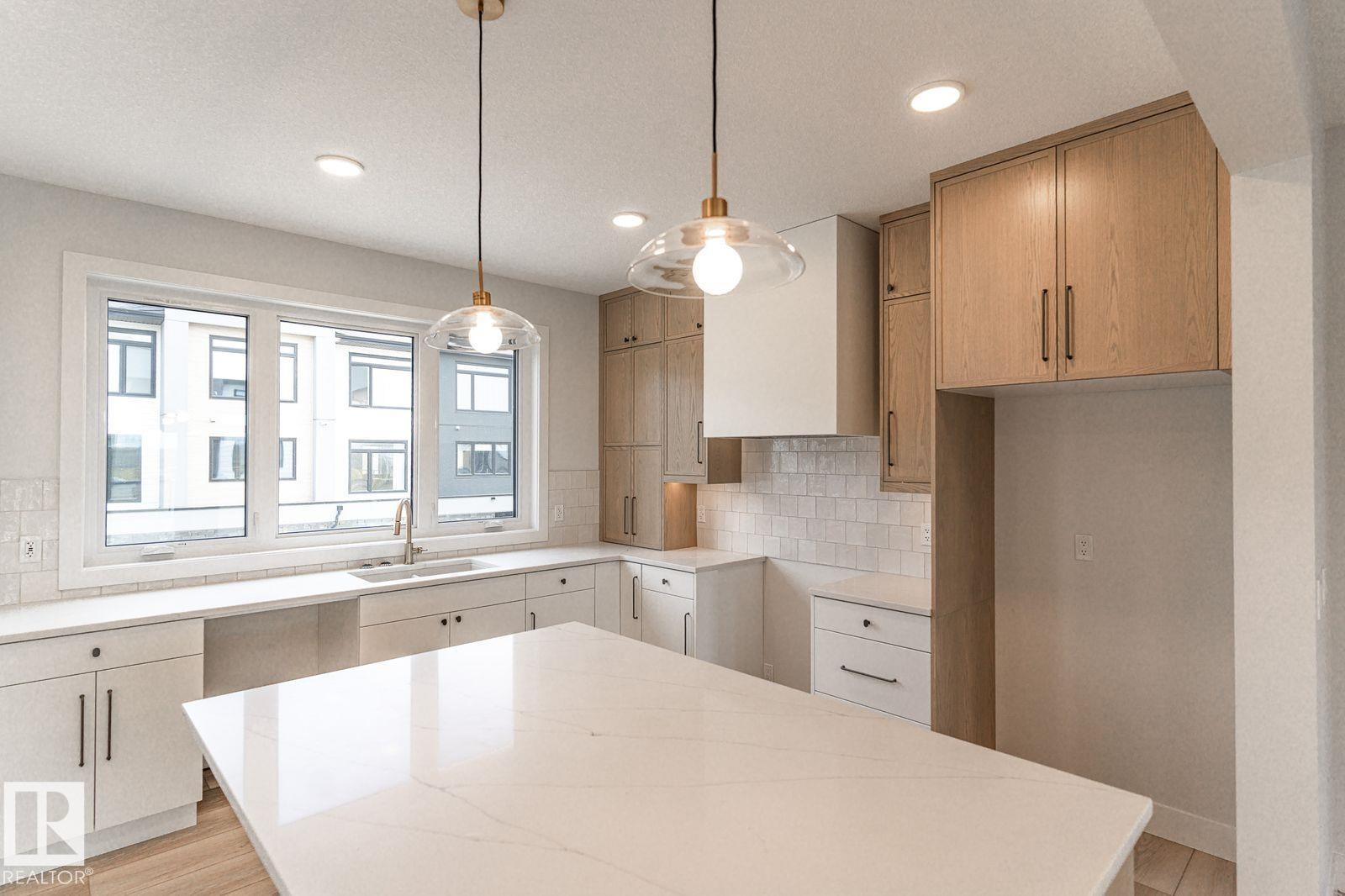 Modern kitchen featuring an expansive island with a white countertop, two pendant lights, recessed lighting, and a window above the sink - 53 7121 May Crescent, Edmonton, AB - Indoor Photo Showing Kitchen With Upgraded Kitchen