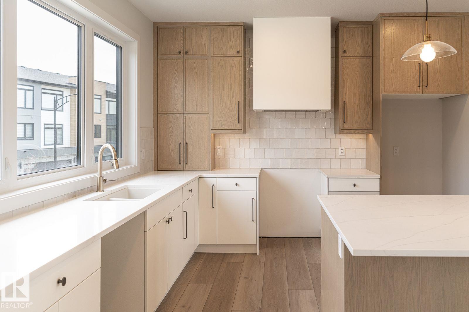 Modern kitchen featuring a large window, light wood-finish and white cabinetry, quartz countertops, a tile backsplash, and wood-finish flooring - 53 7121 May Crescent, Edmonton, AB - Indoor Photo Showing Kitchen With Double Sink