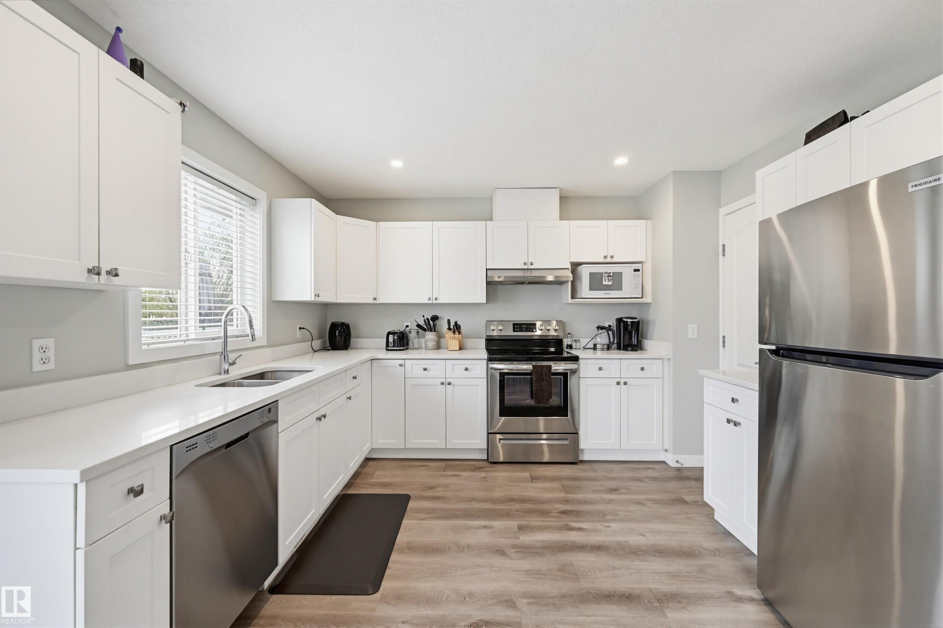 Modern kitchen featuring white shaker cabinetry, stainless steel appliances, light countertops, wood-finish flooring, and recessed lighting - 11 3380 28A Avenue Nw, Edmonton, AB - Indoor Photo Showing Kitchen With Stainless Steel Kitchen With Double Sink