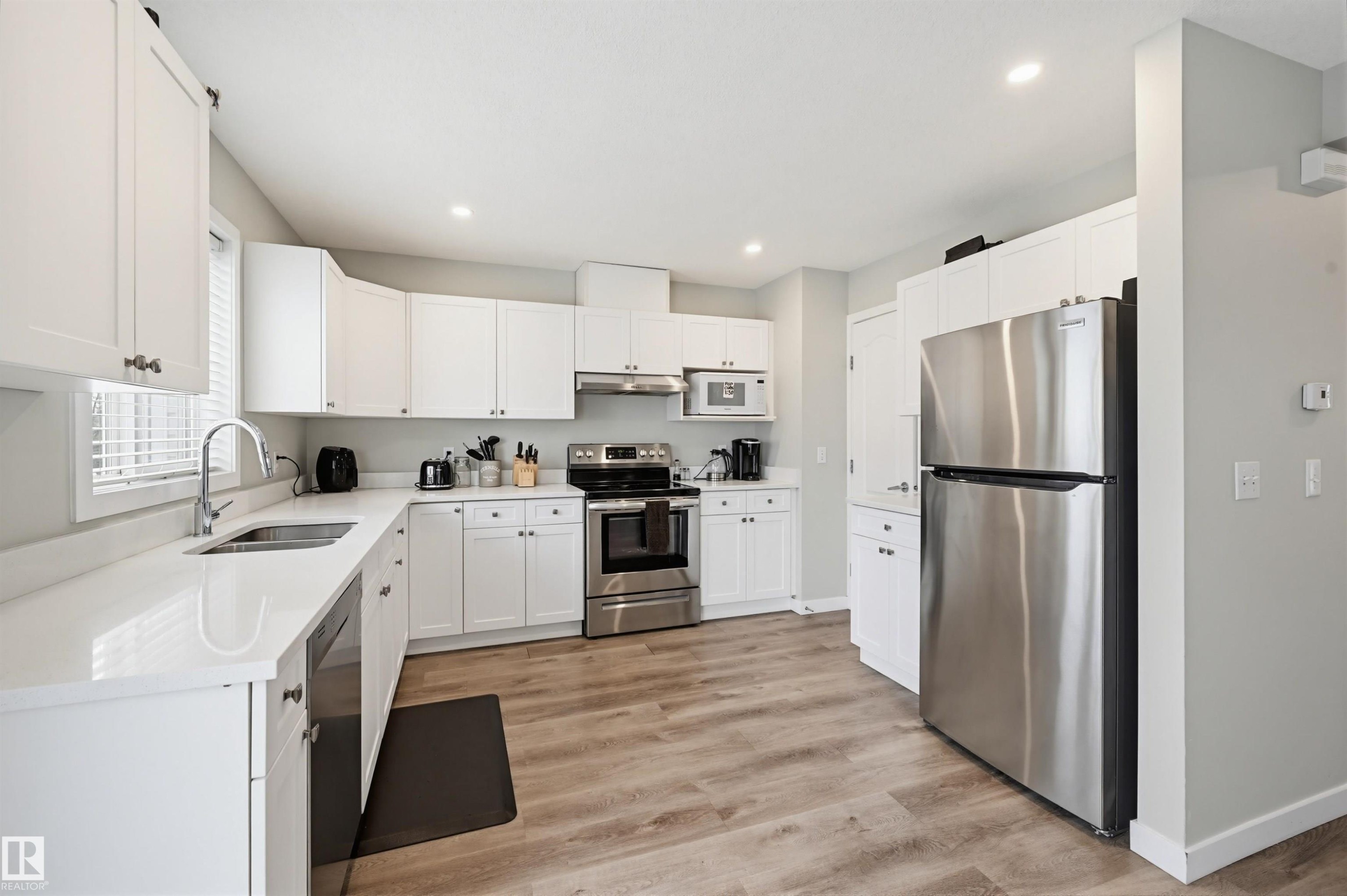 Contemporary kitchen featuring white cabinetry, stainless steel appliances, light-toned wood-finish flooring, recessed lighting, and a window with blinds - 11 3380 28A Avenue Nw, Edmonton, AB - Indoor Photo Showing Kitchen With Stainless Steel Kitchen