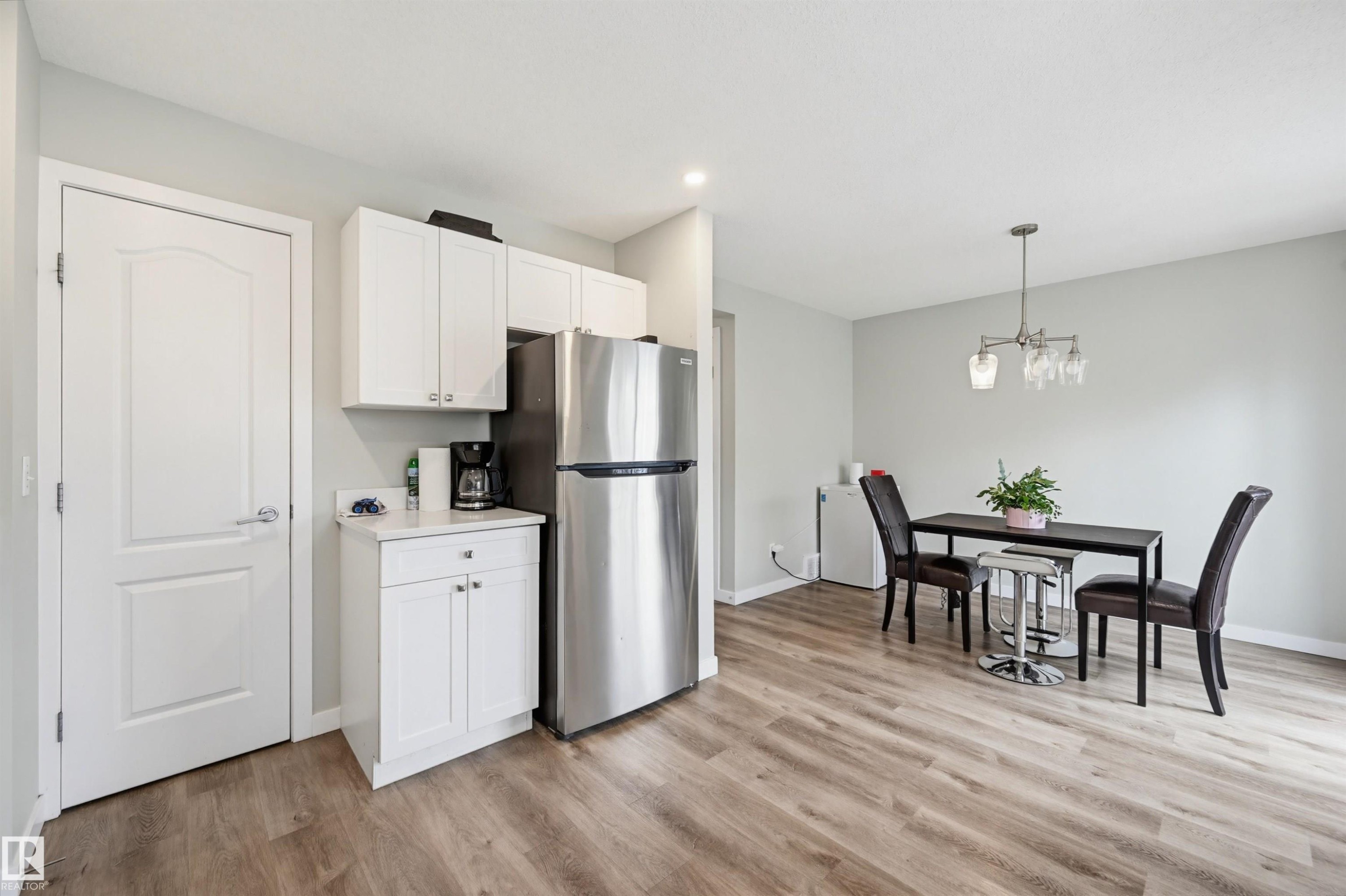 Open-concept kitchen and dining area featuring wood-finish flooring, white cabinetry, a stainless steel refrigerator, and a contemporary light fixture - 11 3380 28A Avenue Nw, Edmonton, AB - Indoor Photo Showing Dining Room
