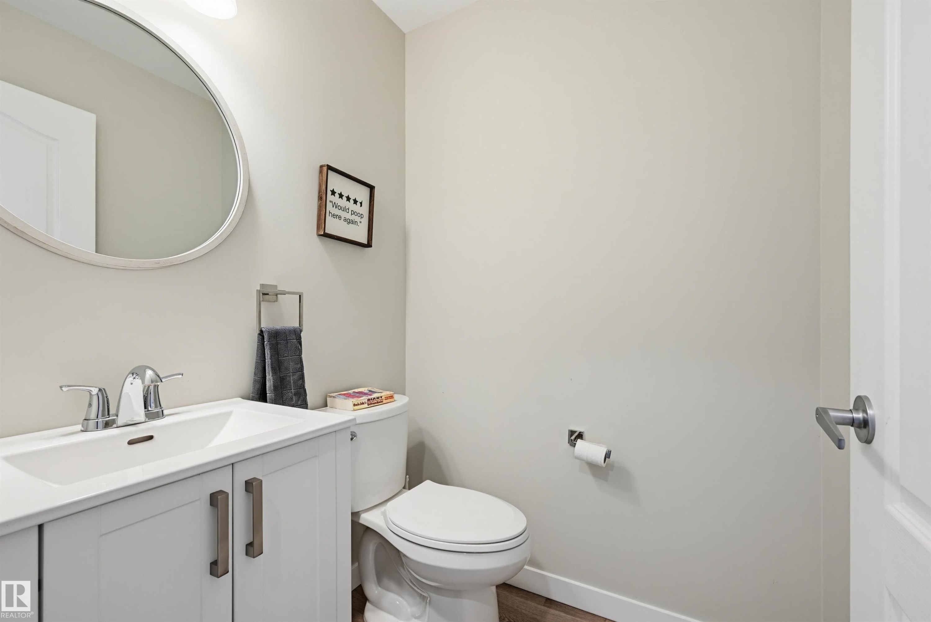 Powder room featuring a white single-sink vanity with chrome faucet, a circular wall-mounted mirror, and wood-finish flooring - 11 3380 28A Avenue Nw, Edmonton, AB - Indoor Photo Showing Other Room