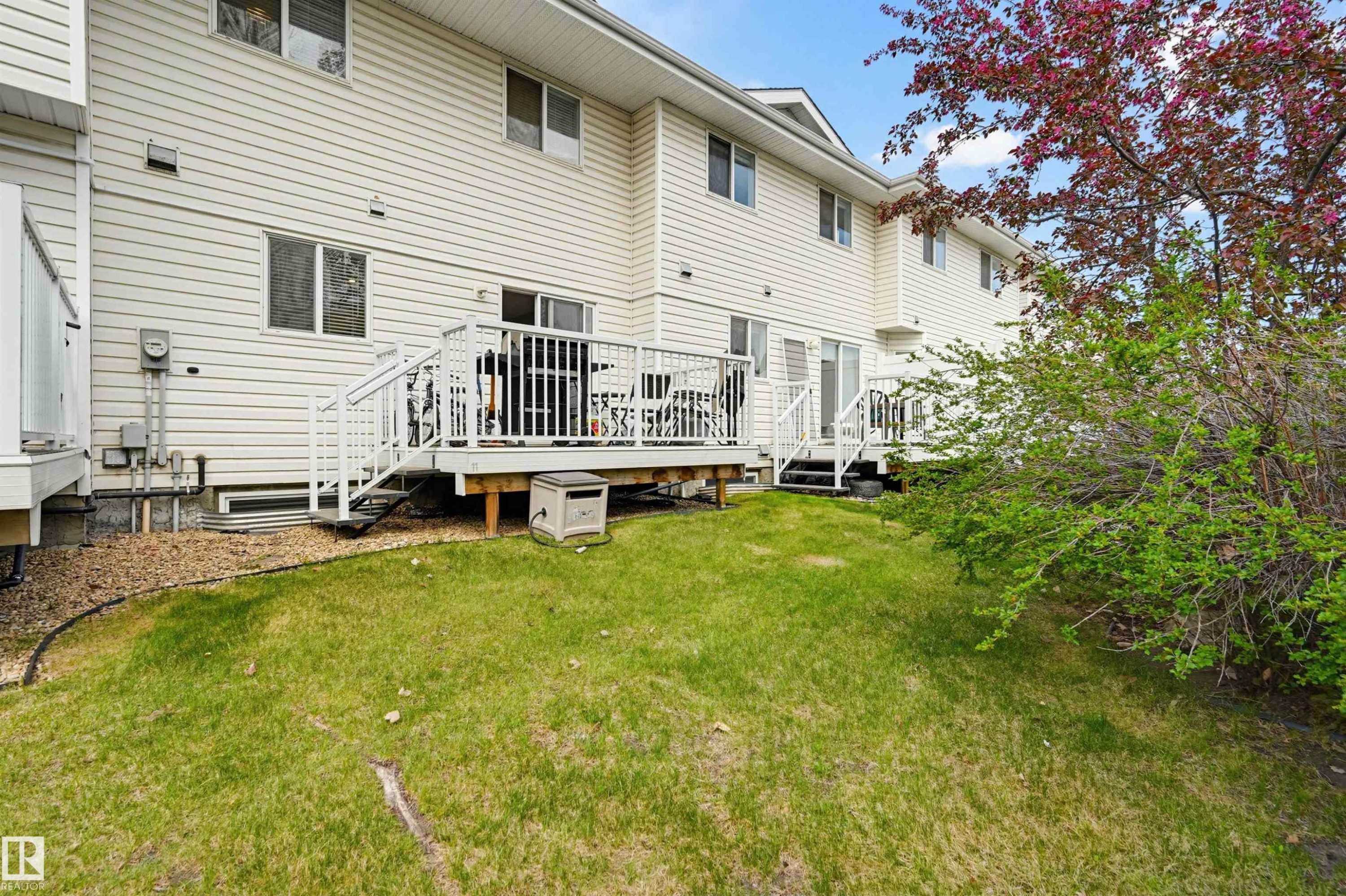 Rear exterior featuring two elevated wooden decks with white railings and stairs, white vinyl siding, multiple windows, and a grassy backyard with mature landscaping - 11 3380 28A Avenue Nw, Edmonton, AB - Indoor