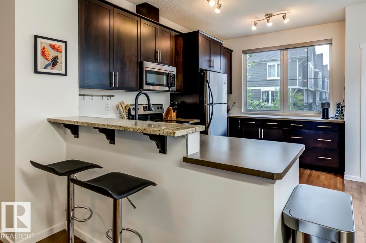 Modern kitchen featuring dark wood-finish cabinetry, stainless steel appliances, a granite breakfast bar, and wood-finish flooring - 67 603 Watt Boulevard, Edmonton, AB - Indoor Photo Showing Kitchen
