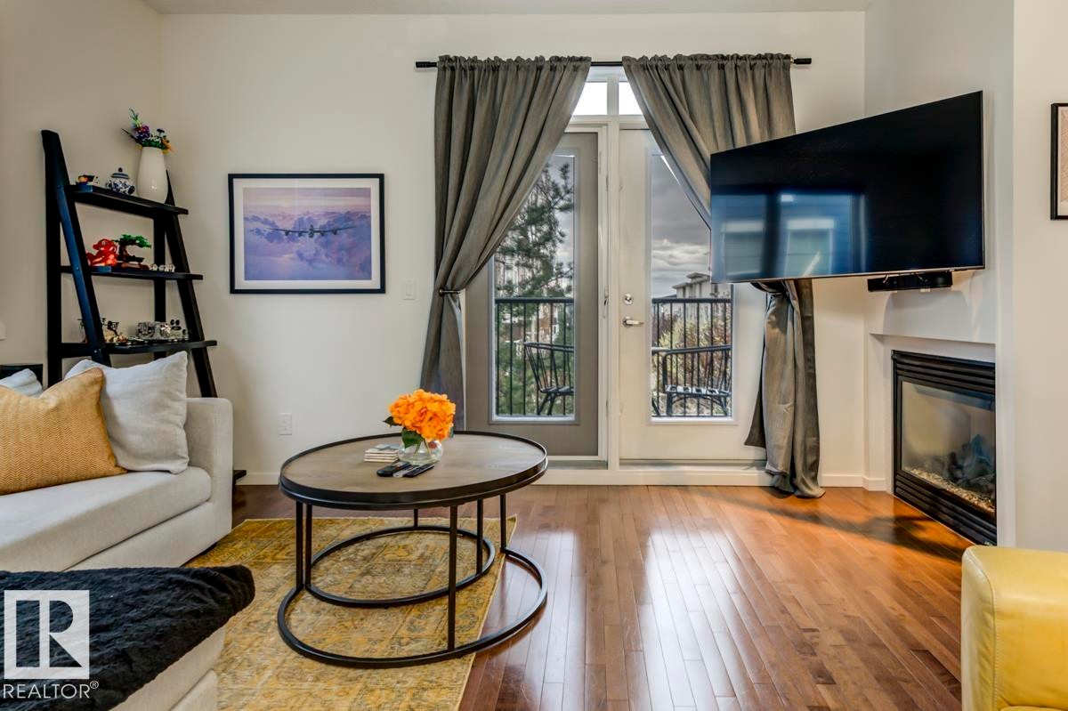 Living area featuring wood-finish flooring, a built-in fireplace, and a glass-paned door leading to an outdoor balcony with a metal railing - 67 603 Watt Boulevard, Edmonton, AB - Indoor Photo Showing Living Room With Fireplace