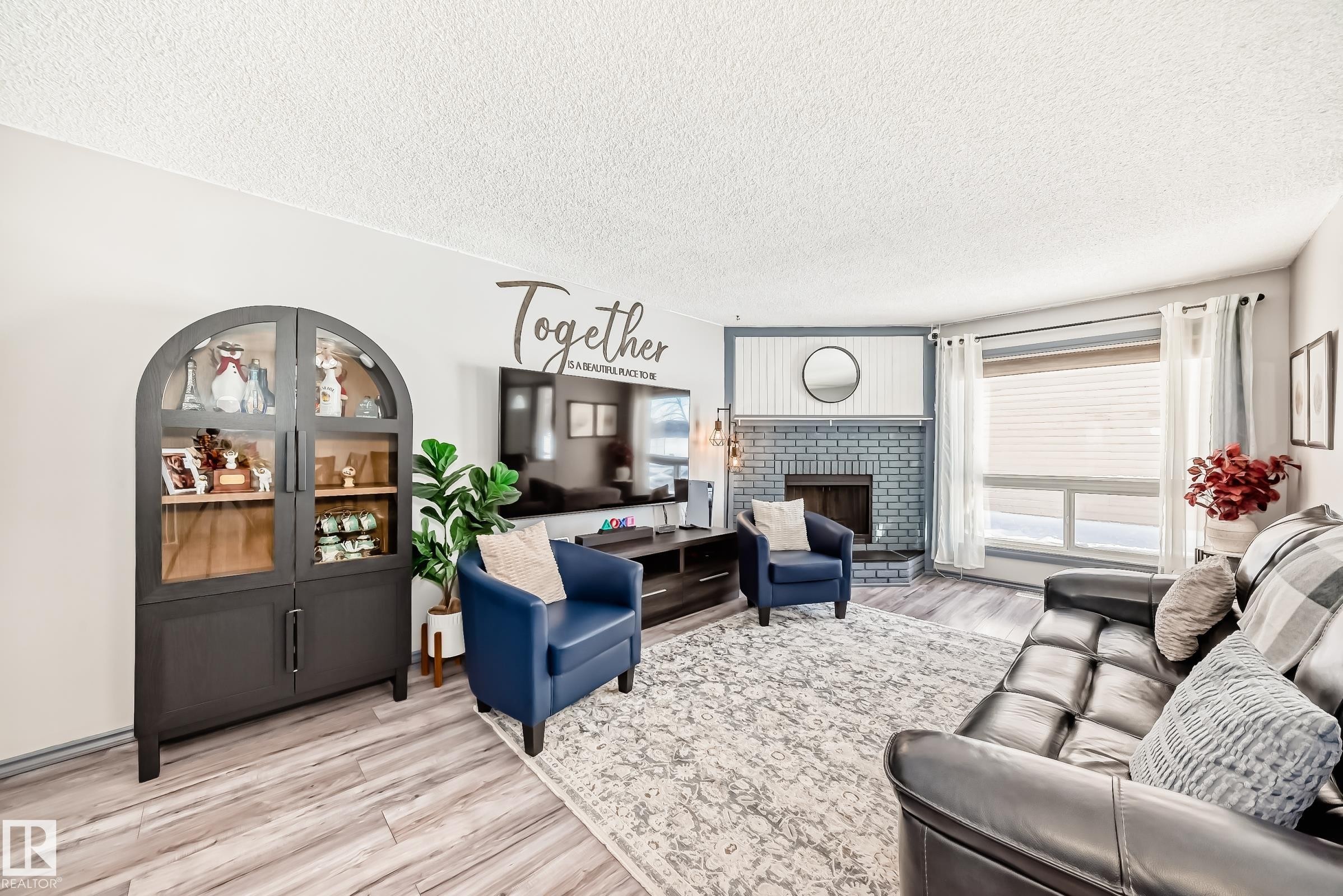 Living area featuring wood-finish flooring, a brick fireplace with a white mantel, and a large window with blinds - 214A Warwick Road, Edmonton, AB - Indoor Photo Showing Living Room With Fireplace