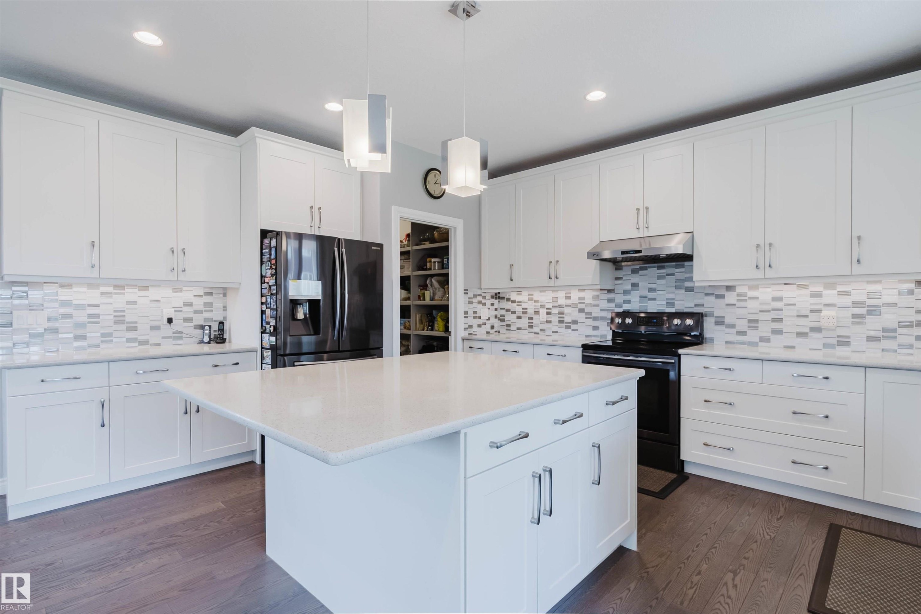 Spacious kitchen featuring an expansive central island with a light-toned countertop, white cabinetry with silver-tone hardware, and wood-finish flooring - 3571 Keswick Boulevard, Edmonton, AB - Indoor Photo Showing Kitchen With Upgraded Kitchen