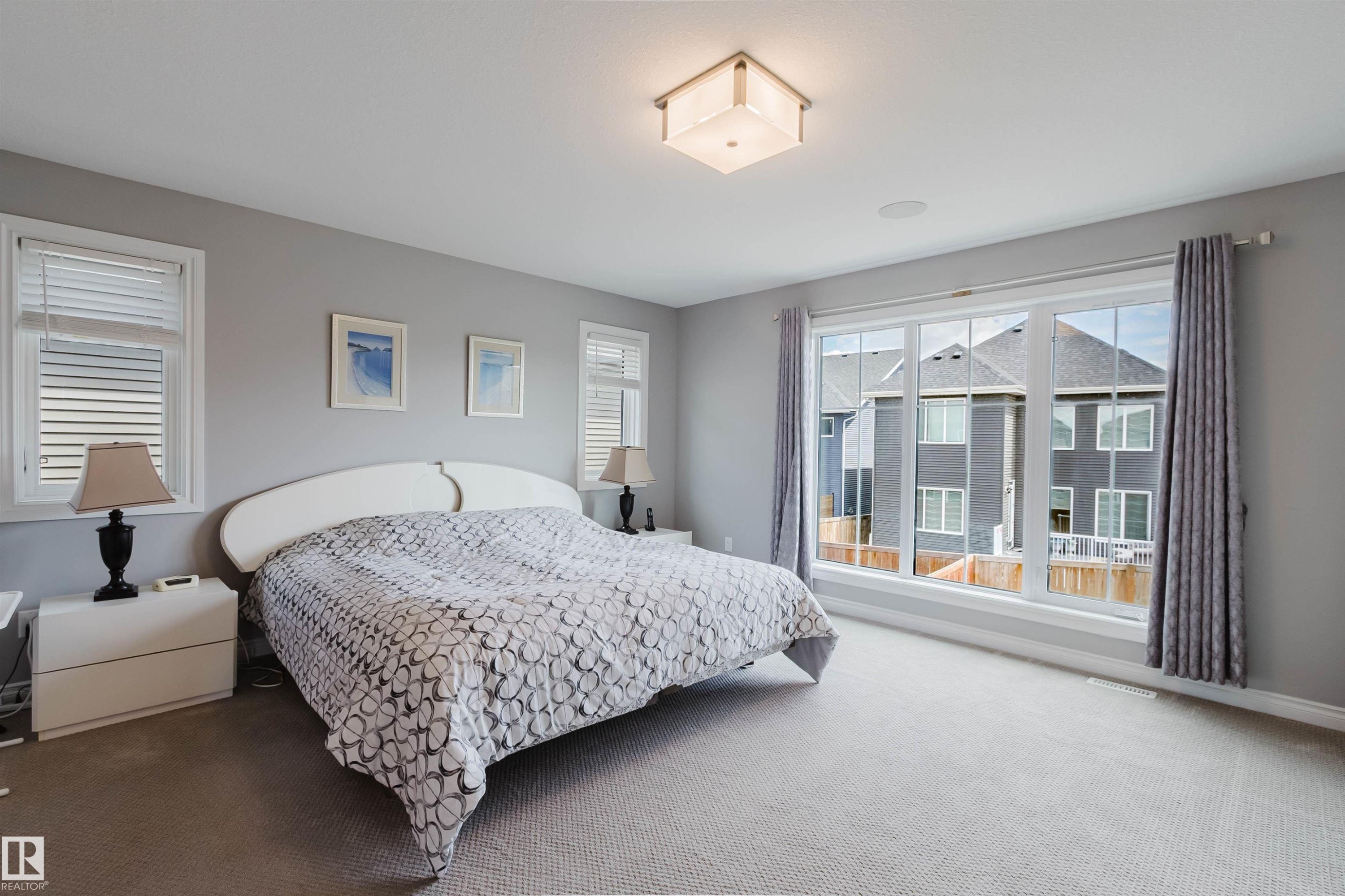 Carpeted bedroom featuring a large picture window, two additional windows, a flush-mount ceiling light fixture, and an integrated ceiling speaker - 3571 Keswick Boulevard, Edmonton, AB - Indoor Photo Showing Bedroom