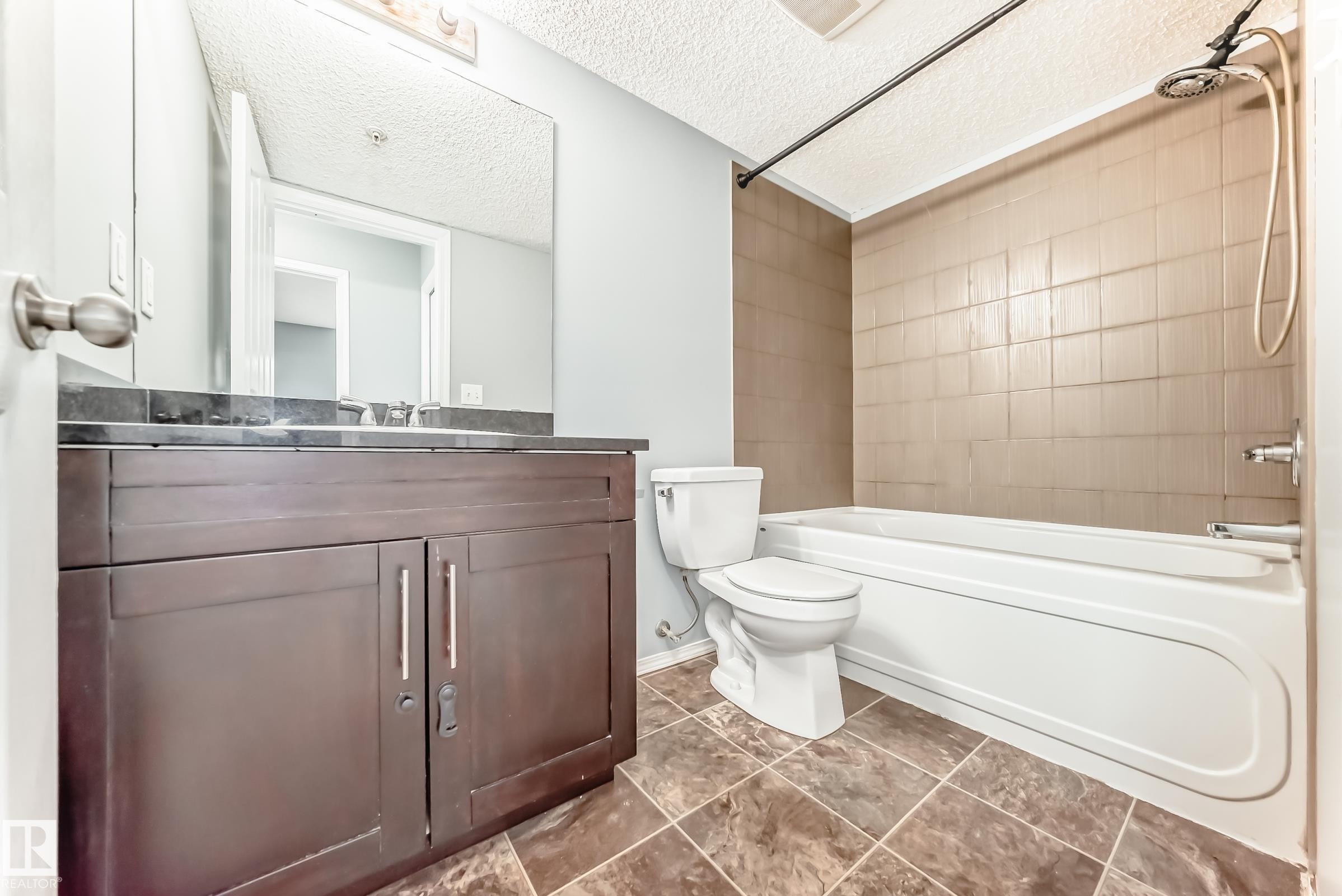 The bathroom features a vanity with a dark countertop and a white sink, a toilet, a bathtub with a shower, and tiled flooring - 101 530 Watt Boulevard, Edmonton, AB - Indoor Photo Showing Bathroom