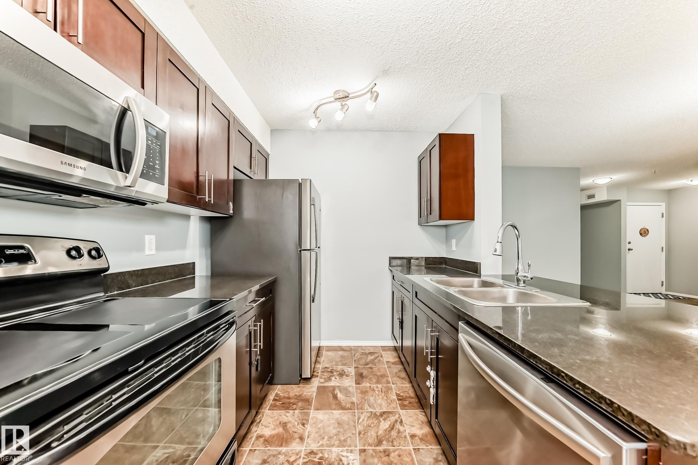 The kitchen features dark wood cabinetry, stainless steel appliances, a double basin sink, and tiled flooring - 101 530 Watt Boulevard, Edmonton, AB - Indoor Photo Showing Kitchen With Double Sink
