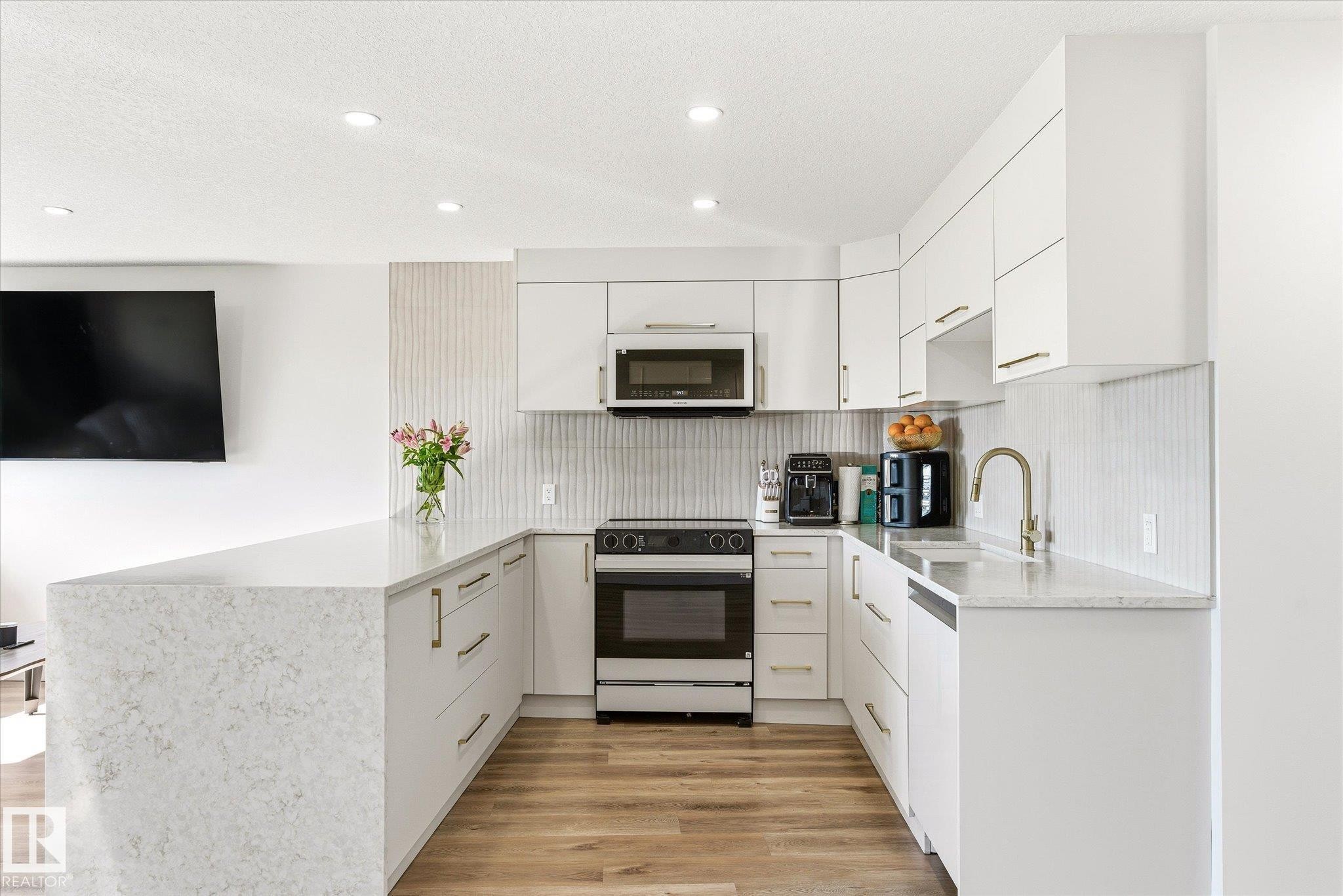 Modern kitchen featuring white cabinetry with gold-tone hardware, a U-shaped countertop with a waterfall edge, stainless steel appliances, a white vertical tile backsplash, and wood-finish flooring - 108 10105 95 Street, Edmonton, AB - Indoor Photo Showing Kitchen With Upgraded Kitchen