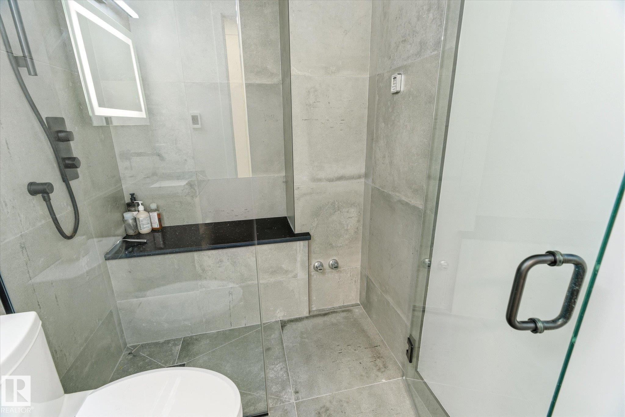 Contemporary shower enclosure featuring full-height glass panels, matte black shower fixtures, a built-in bench with a dark countertop, and large format gray tile surrounds - 108 10105 95 Street, Edmonton, AB - Indoor Photo Showing Bathroom