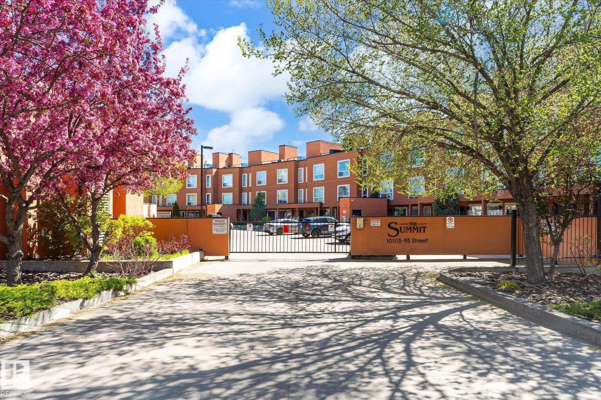 Gated community entrance with mature landscaping, featuring a vibrant pink flowering tree and green foliage - 108 10105 95 Street, Edmonton, AB - Outdoor