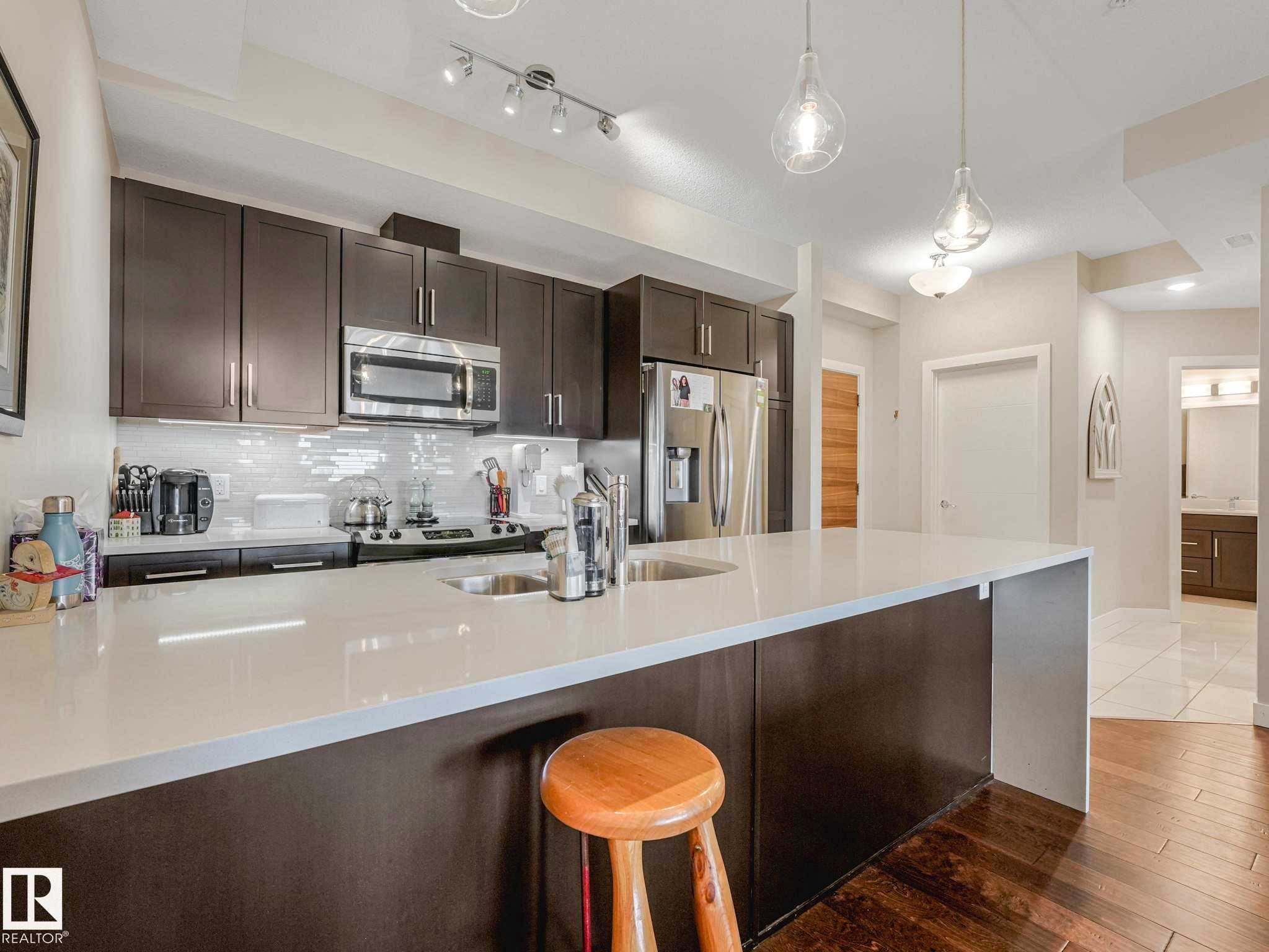 Modern kitchen with a large central island, white countertop, dark wood-finish cabinetry, stainless steel appliances, and wood-finish flooring - 213 625 Leger Way, Edmonton, AB - Indoor Photo Showing Kitchen With Stainless Steel Kitchen With Double Sink With Upgraded Kitchen