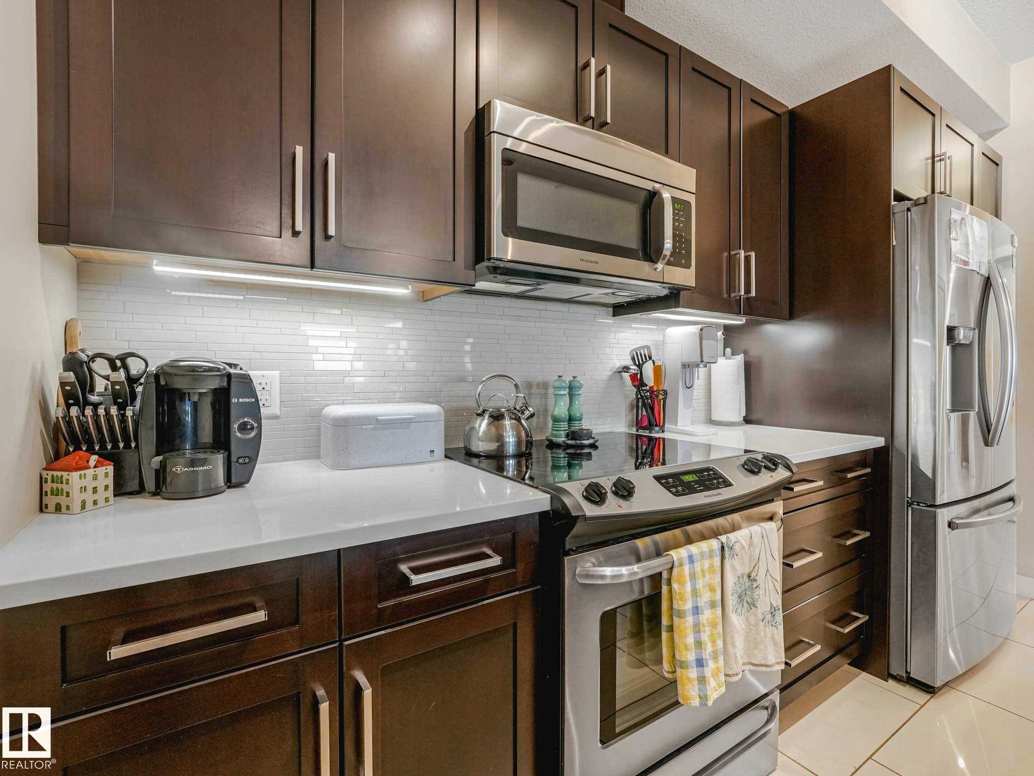 Modern kitchen featuring dark wood-finish cabinetry with brushed nickel hardware, white countertops, and a white subway tile backsplash - 213 625 Leger Way, Edmonton, AB - Indoor Photo Showing Kitchen