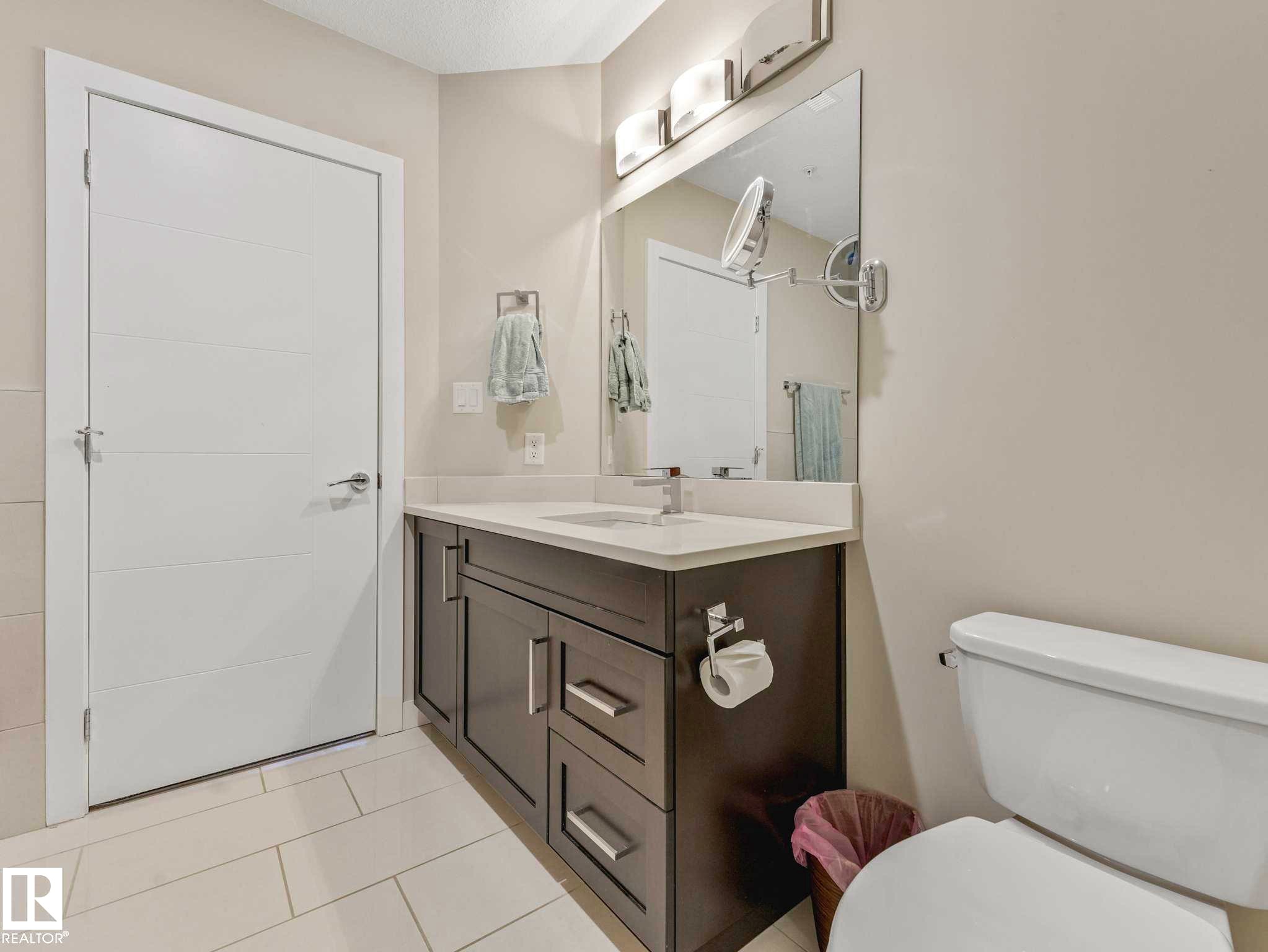Bathroom featuring a dark wood-finish vanity with a light countertop, integrated sink, and an expansive mirror - 213 625 Leger Way, Edmonton, AB - Indoor Photo Showing Bathroom