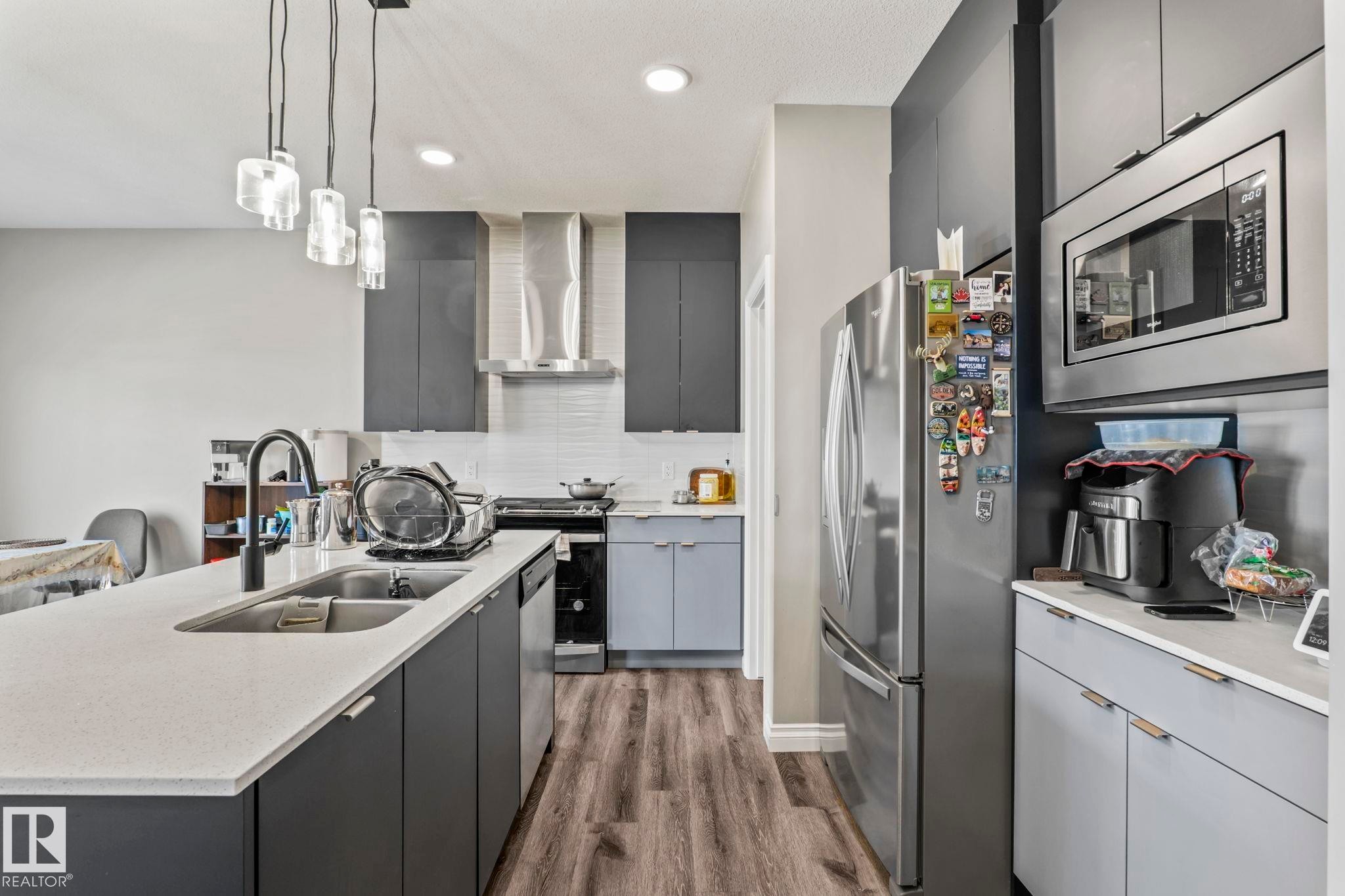 Modern kitchen featuring a spacious island with an undermount sink, pendant lighting, stainless steel appliances, dark cabinetry, and wood-finish flooring - 5142 23A Avenue, Edmonton, AB - Indoor Photo Showing Kitchen With Double Sink With Upgraded Kitchen