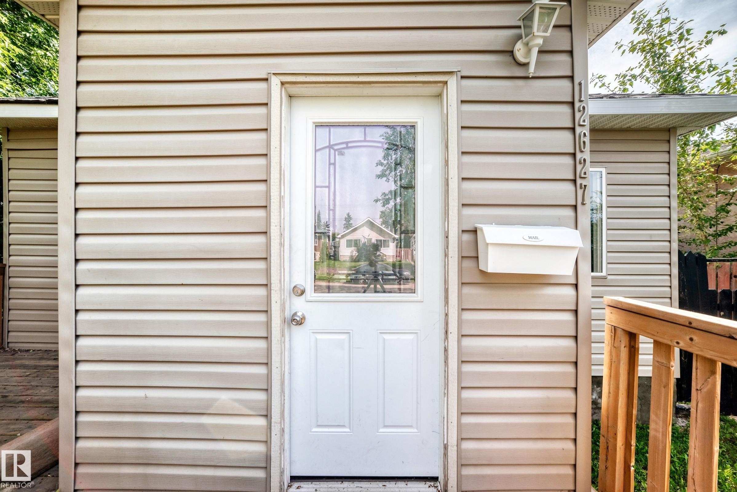 Front entrance featuring a white door with decorative glass, beige horizontal siding, and a wall-mounted light fixture - 12627 71 Street Nw, Edmonton, AB
