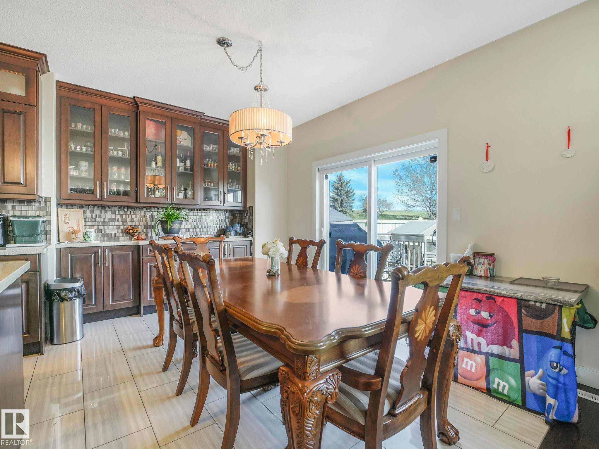 Dining area featuring a wood-finish dining table, drum chandelier, built-in cabinetry with glass fronts, a tile backsplash, and sliding glass doors - 3910 49 Avenue, Beaumont, AB - Indoor Photo Showing Dining Room