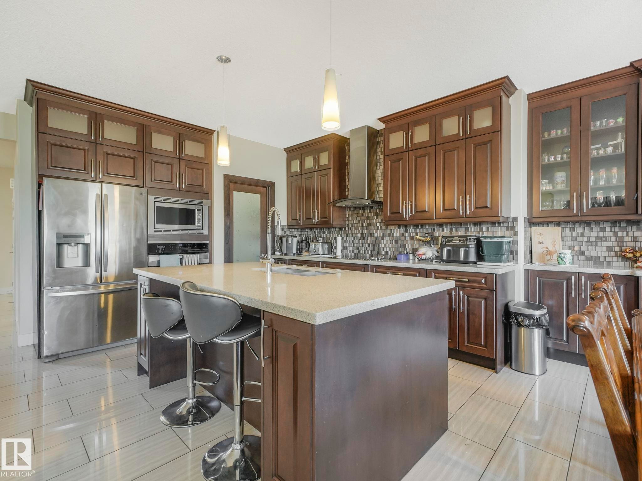 Kitchen featuring an expansive center island, extensive dark wood cabinetry, stainless steel appliances, a mosaic tile backsplash, and light-toned tile flooring - 3910 49 Avenue, Beaumont, AB - Indoor Photo Showing Kitchen With Stainless Steel Kitchen With Upgraded Kitchen