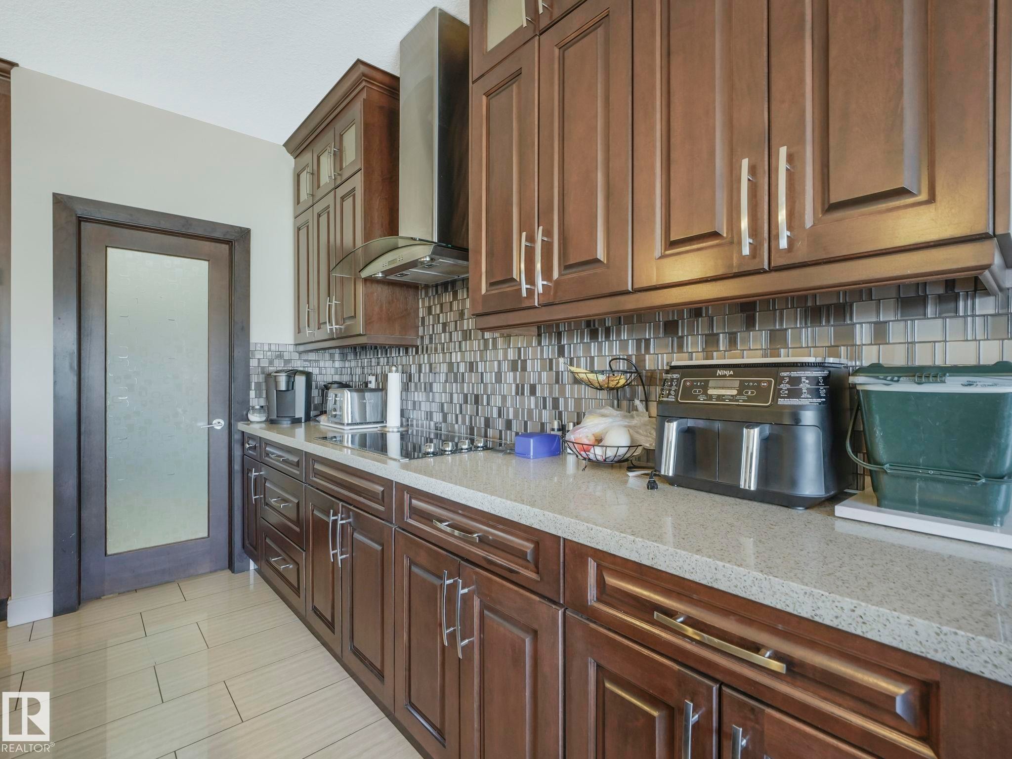 Kitchen featuring rich wood cabinetry, patterned tile backsplash, light-toned countertops, and a modern stainless steel range hood - 3910 49 Avenue, Beaumont, AB - Indoor Photo Showing Kitchen