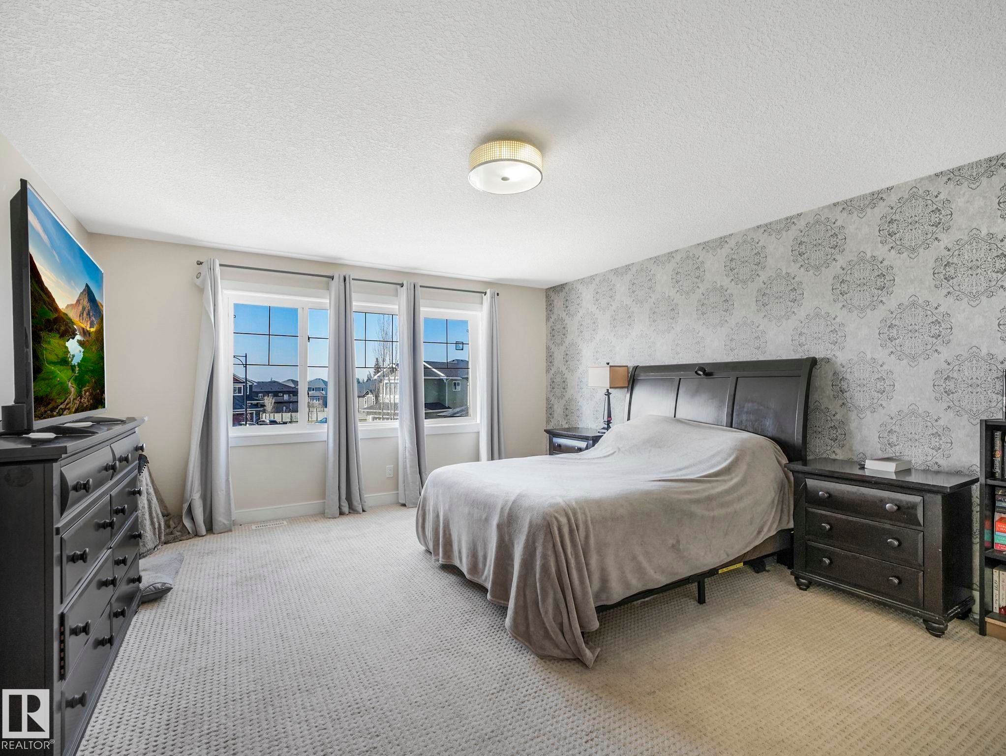 Spacious room featuring patterned accent wall, three large windows, textured carpeting, and a flush mount ceiling light fixture - 3910 49 Avenue, Beaumont, AB - Indoor Photo Showing Bedroom