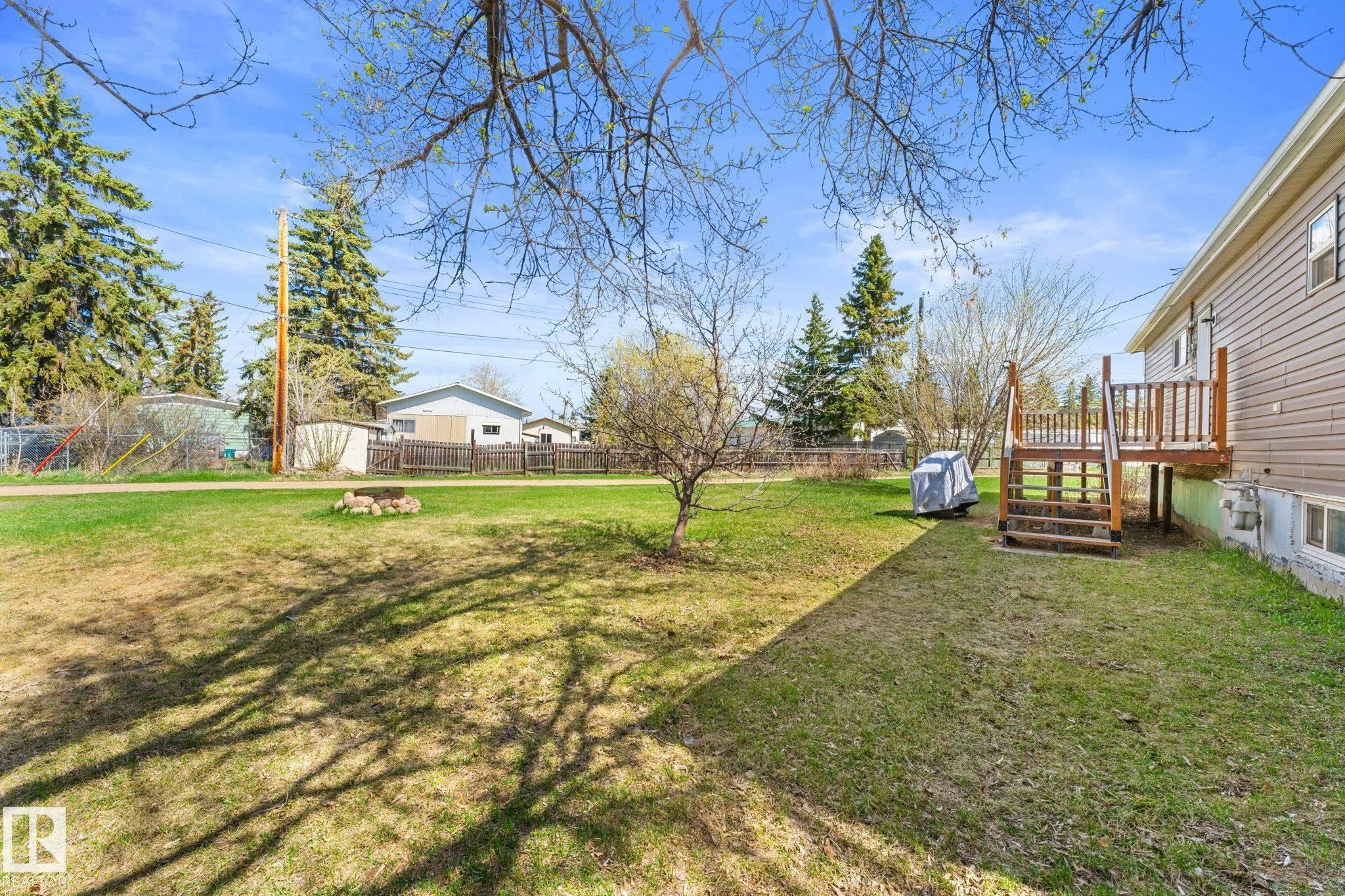 Expansive rear yard featuring a wood deck with stairs, a fire pit, and mature perimeter trees - 4508 50A Avenue, Barrhead, AB - Outdoor